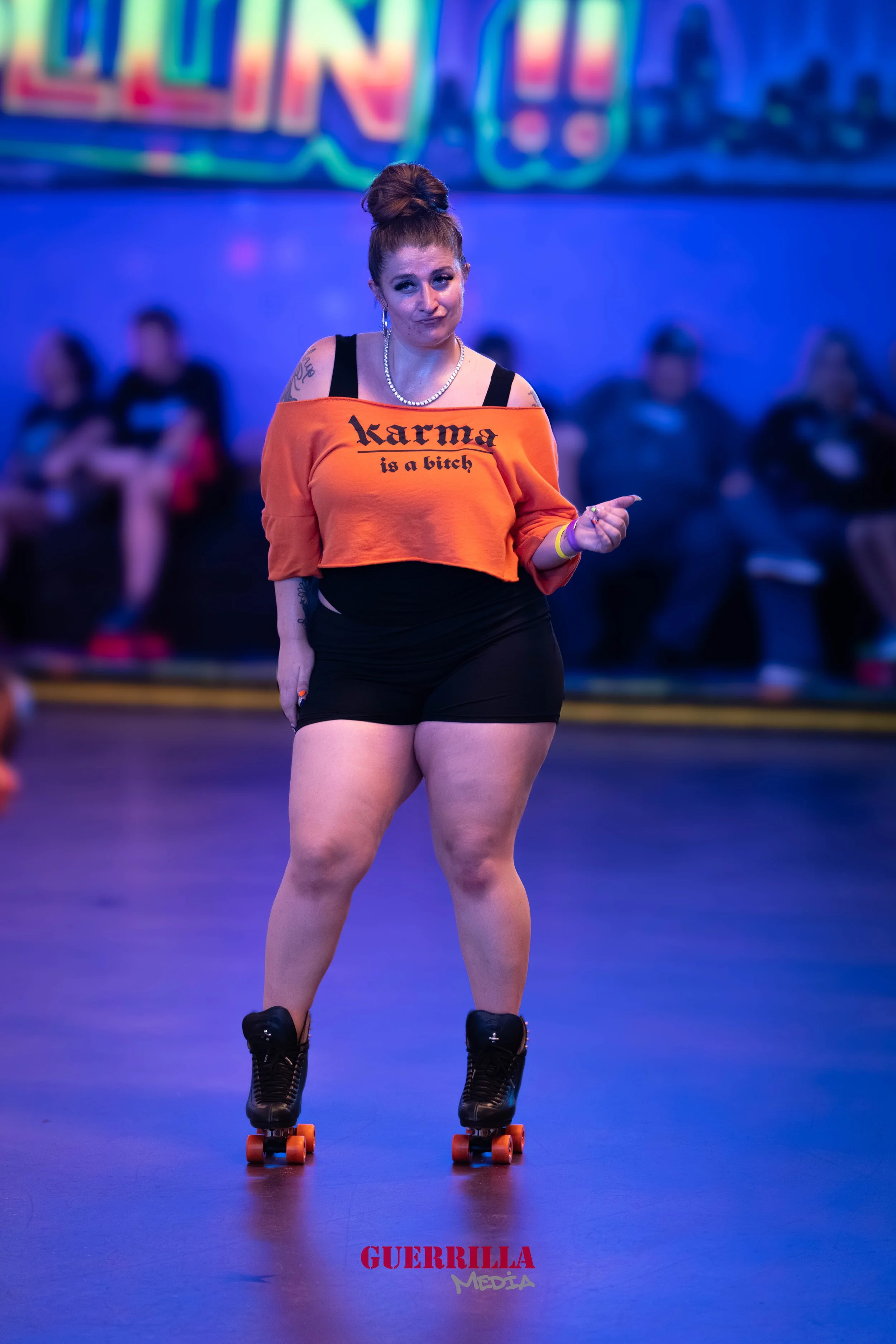 A woman rollerskating indoors, wearing an orange shirt with black text that reads "karma is a bitch," black shorts, and black roller skates with orange wheels. She has her hair in a bun, tattoos, and is making a gesture with her hand. There are blurr