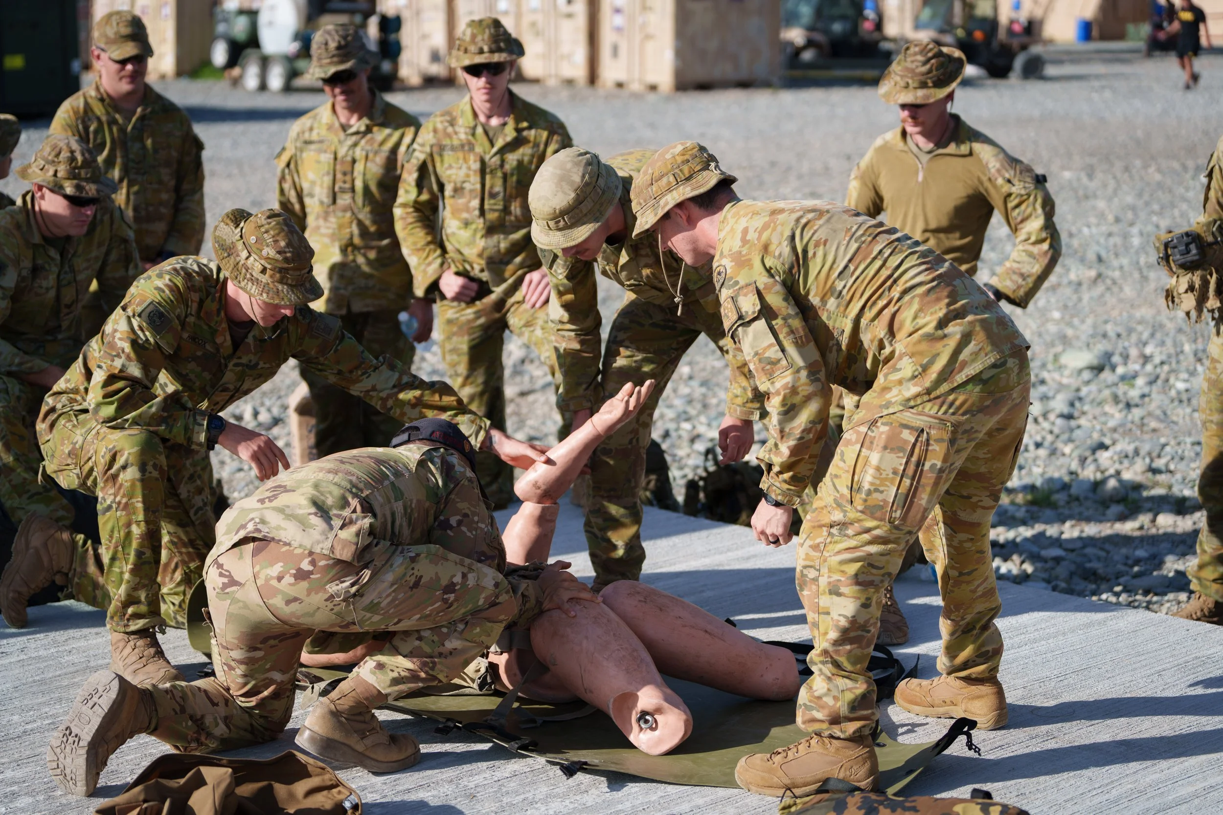 Military personnel in camouflage uniforms practicing a medical or rescue procedure on a training dummy or mannequin outdoors.