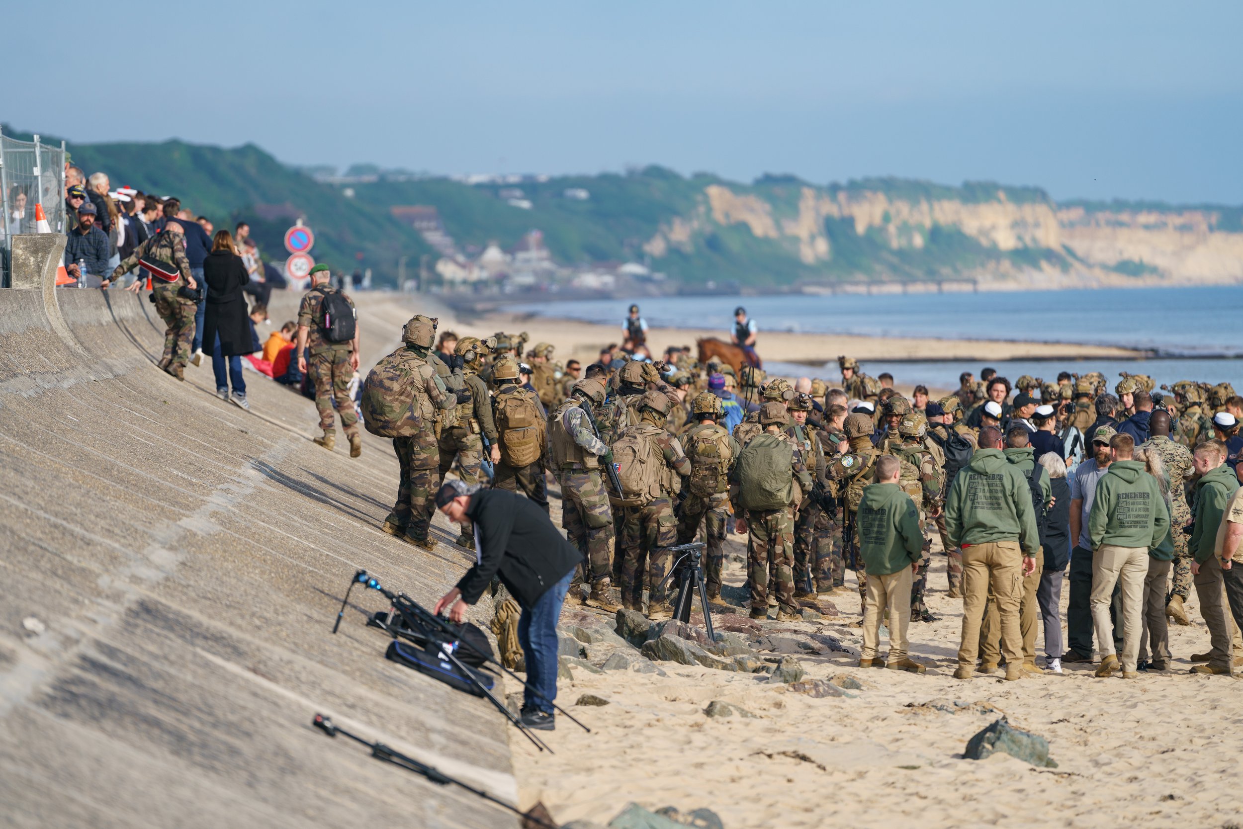 Large crowd of soldiers and civilians gathered on a beach, with some in military uniforms, under a cloudy sky, near cliffs.