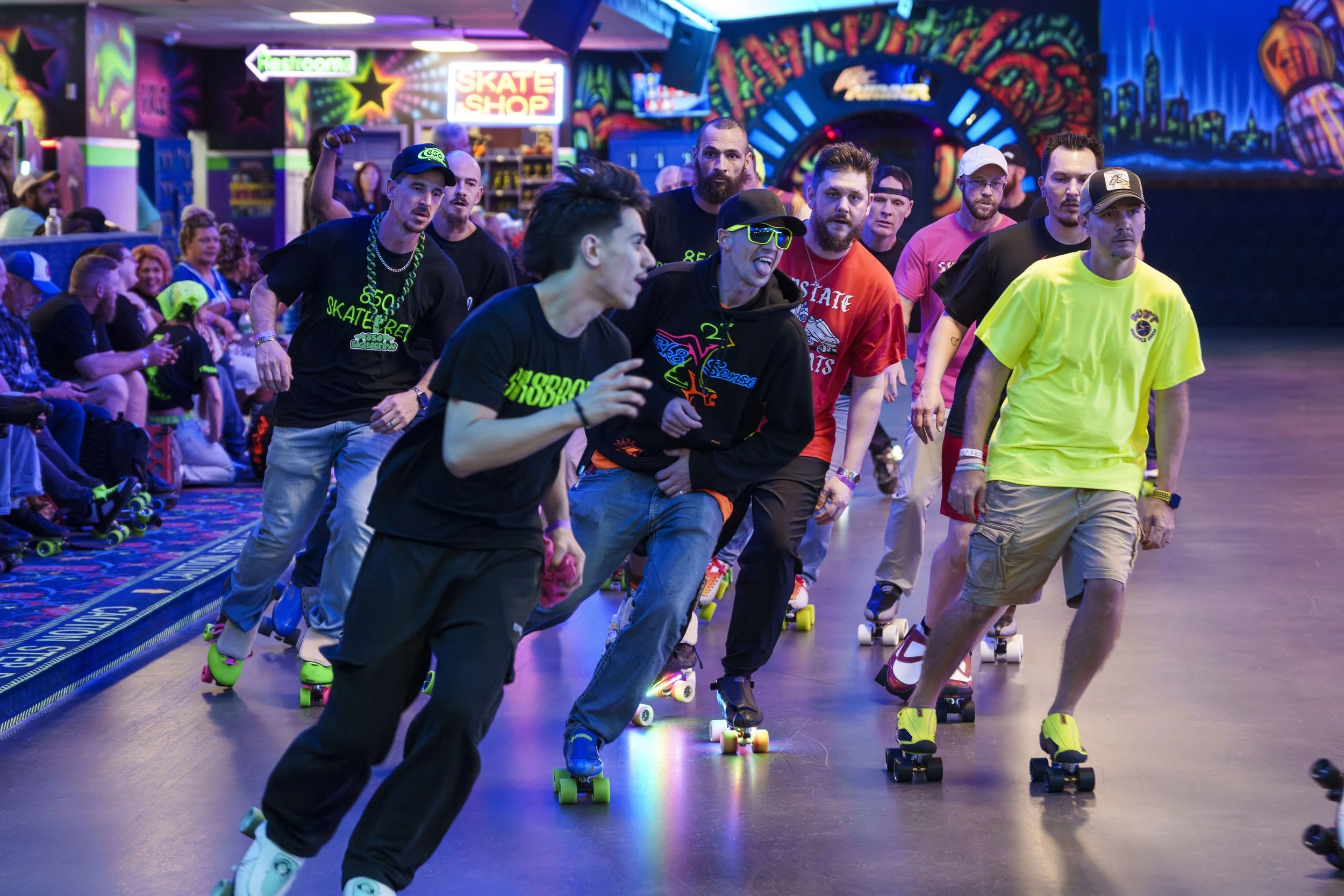 A group of people roller skating indoors at a brightly lit roller rink with colorful neon signs and eye-catching wall murals in the background, including a sign that says 'Skate Shop'.