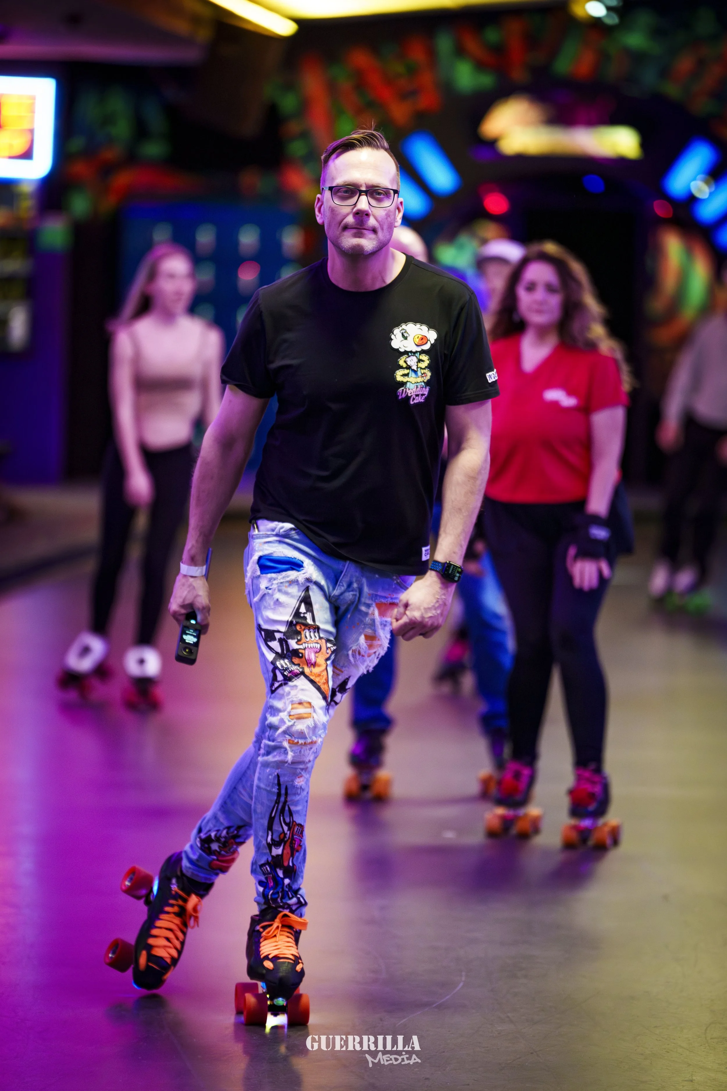 A man roller skating in an indoor roller rink with colorful neon lights, wearing a black T-shirt, ripped jeans, and orange roller skates, holding a small device in his right hand.