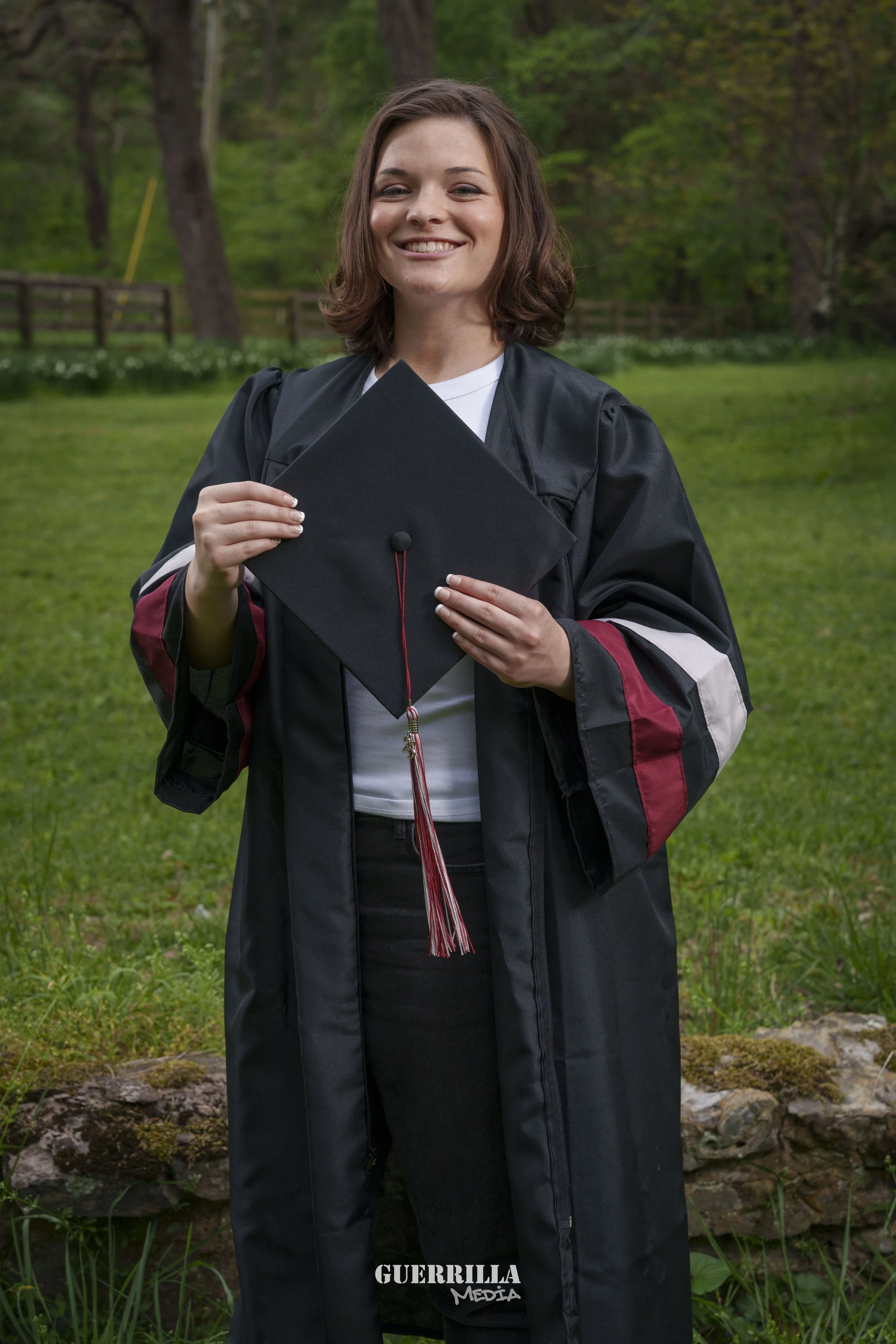 A young woman in a graduation cap and gown holding her cap, standing outdoors on a grassy area with trees in the background, smiling at the camera.