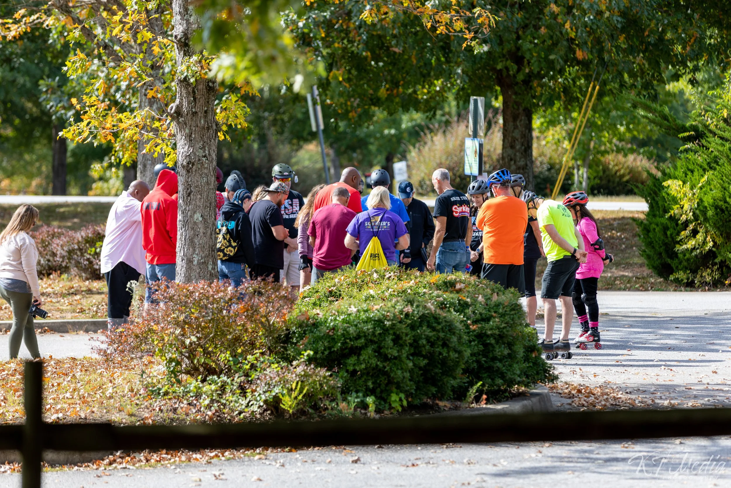 A group of people gathered outdoors on a sidewalk surrounded by trees and bushes, some with helmets and roller skates, participating in an outdoor activity on a sunny day.