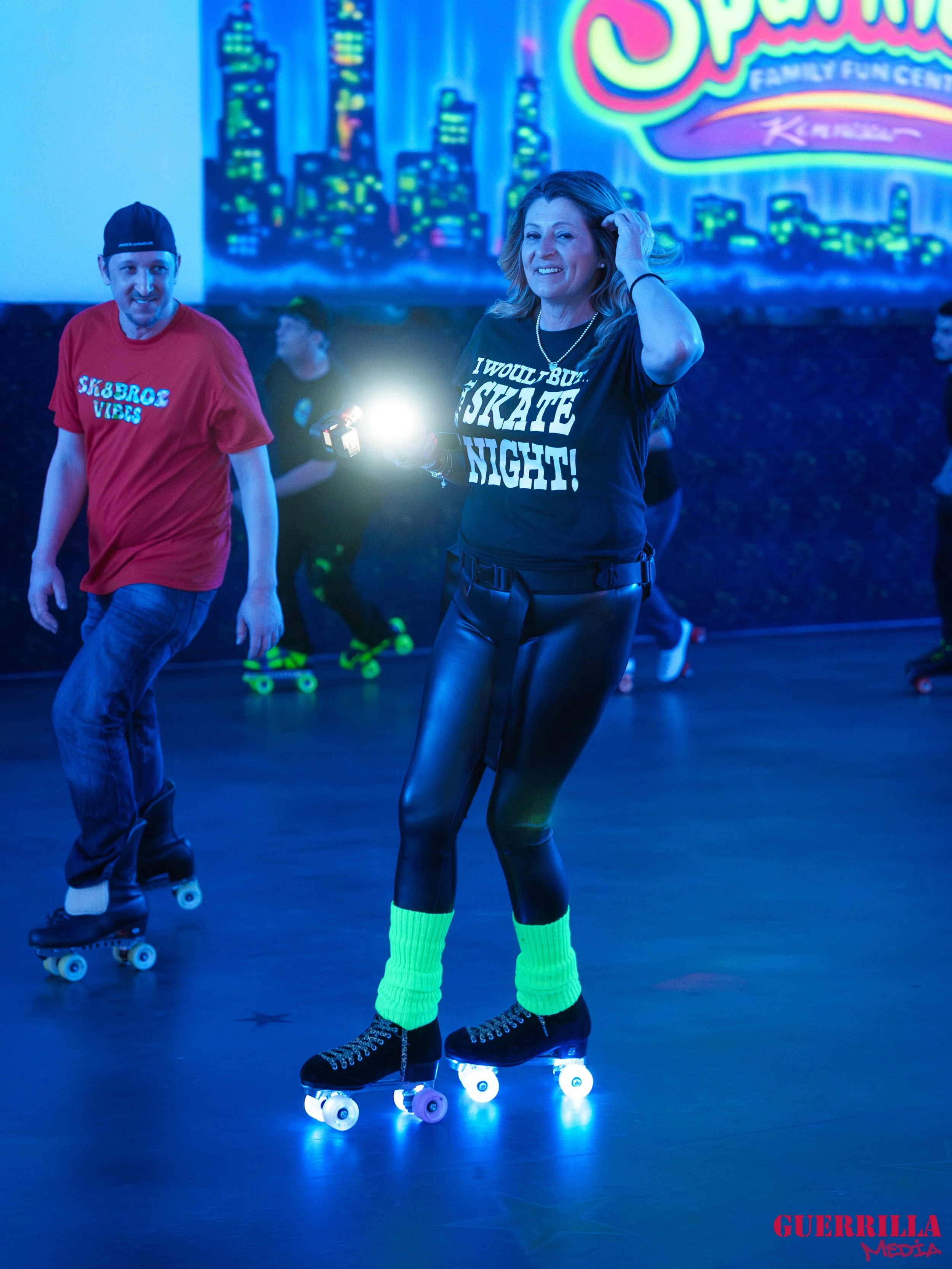 People roller skating indoors with neon lights and a cityscape mural in the background, including a woman smiling and wearing a black t-shirt that says 'I WOULD BUST A SKATE NIGHT!'