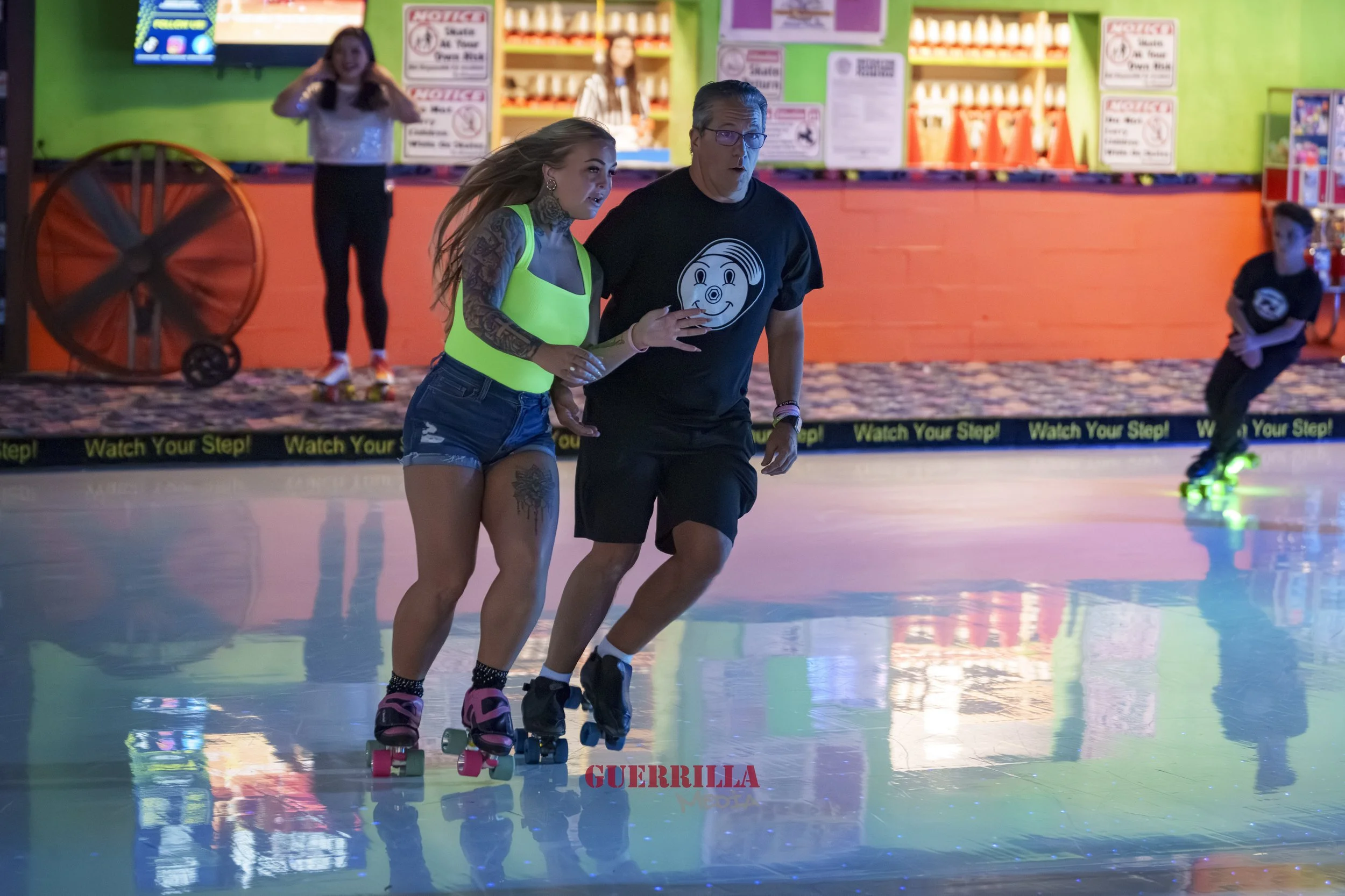 A woman and man roller skating in an indoor rink, with the woman assisting the man. Two people are in the background near the counter, and colorful signage and a fan are visible in the background.