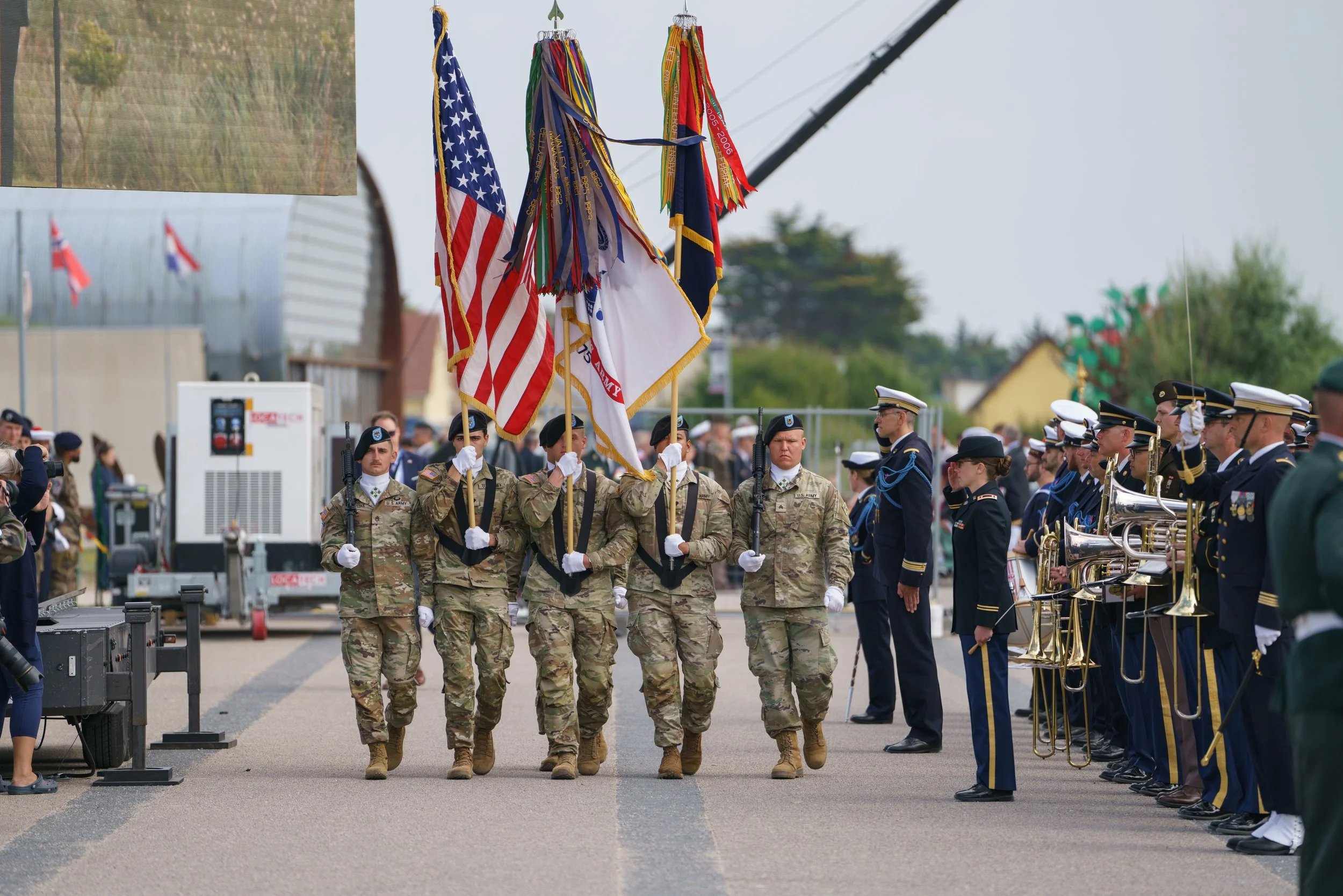 Military personnel in uniform carrying flags, performing a flag procession during a ceremony, with band members standing at attention on the side.