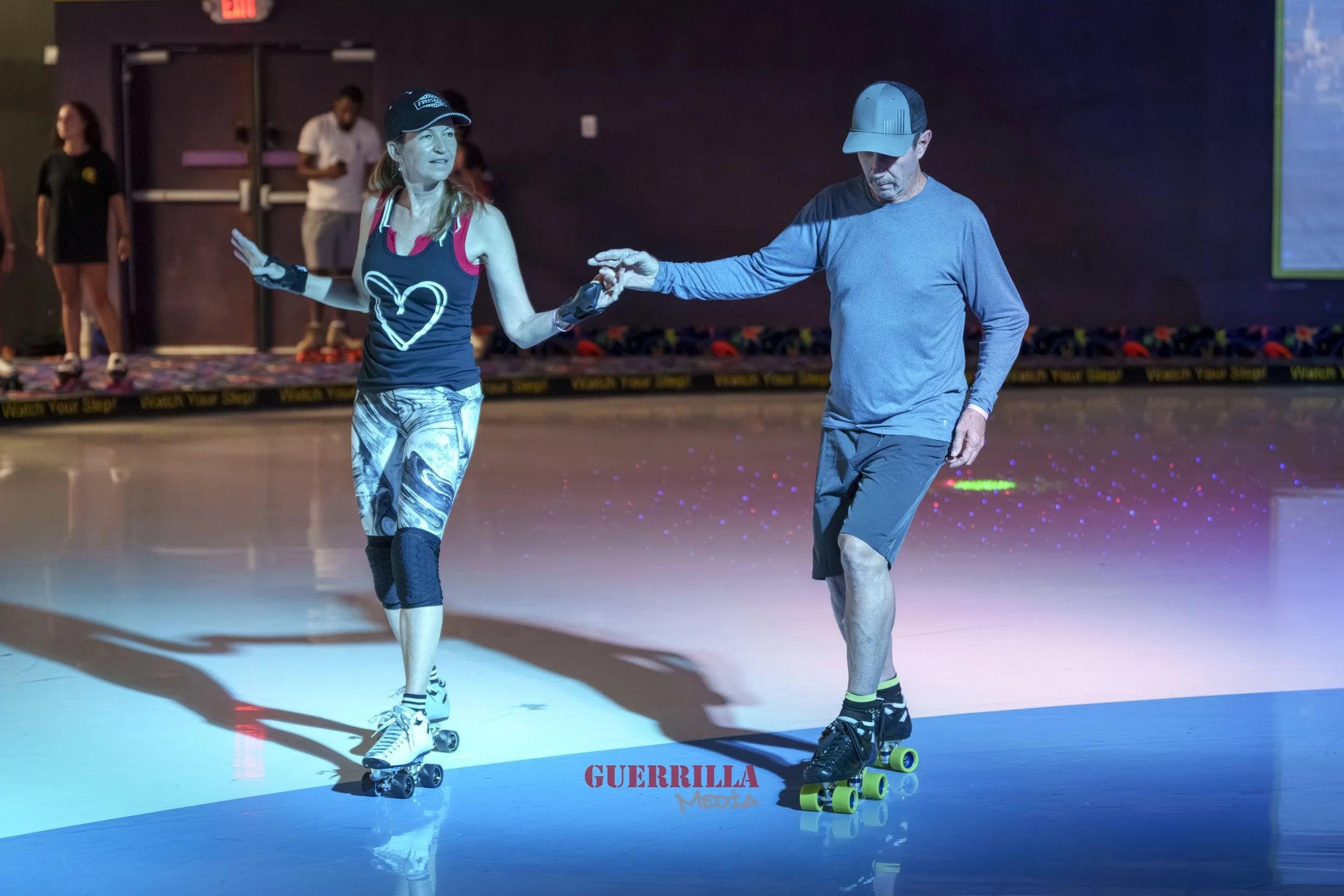 A woman and a man roller skating on an indoor rink. The woman is wearing a black tank top with a heart design, patterned leggings, a cap, and wrist guards. The man is dressed in a gray long-sleeve shirt, gray shorts, and a cap. She is holding his han