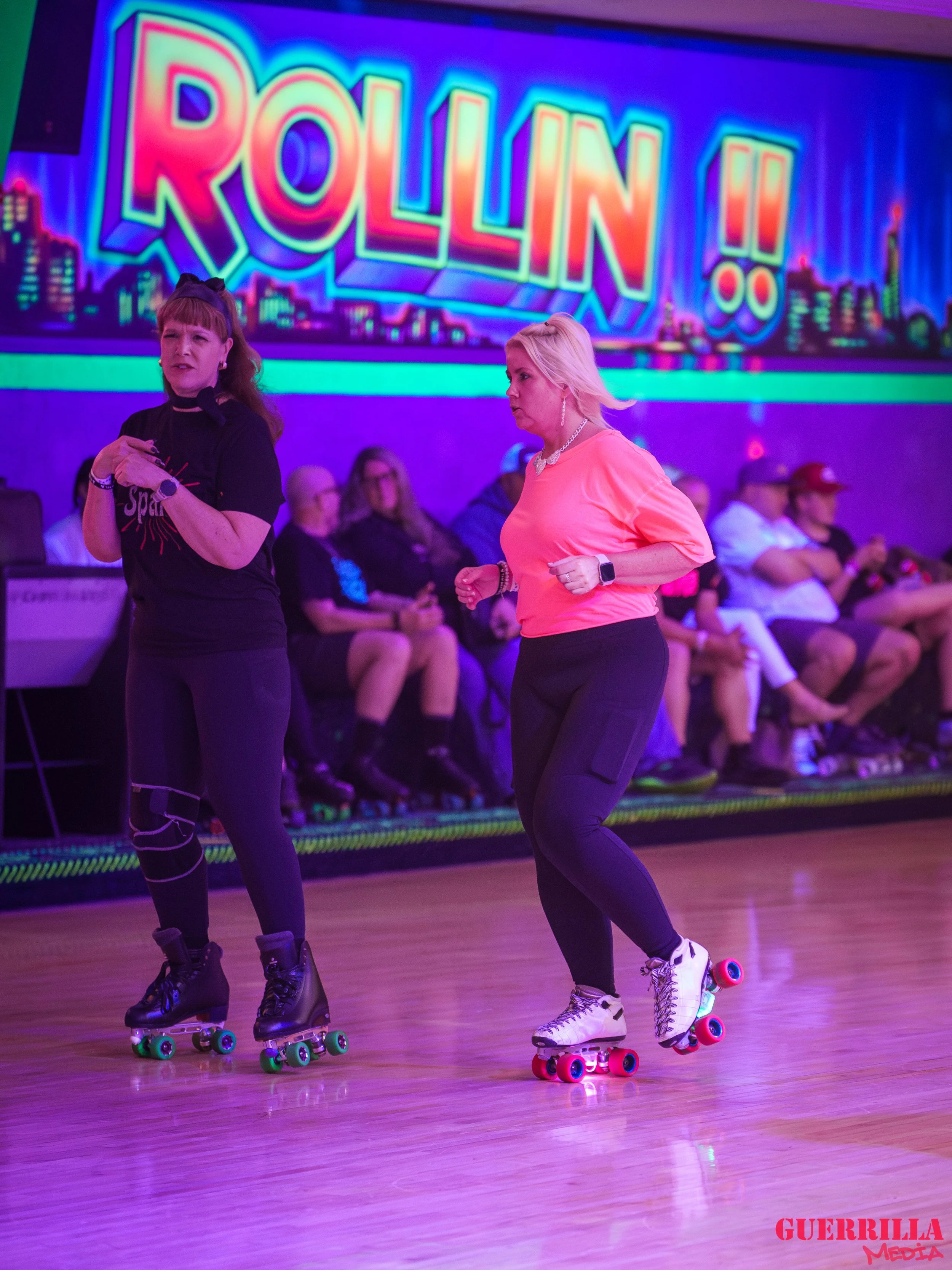 Two women roller skating in an indoor rink with a colorful neon sign that says "ROLLIN!!" in the background. Multiple people are seated along the side, observing. The woman on the left wears a black shirt and black roller skates with green wheels, wh