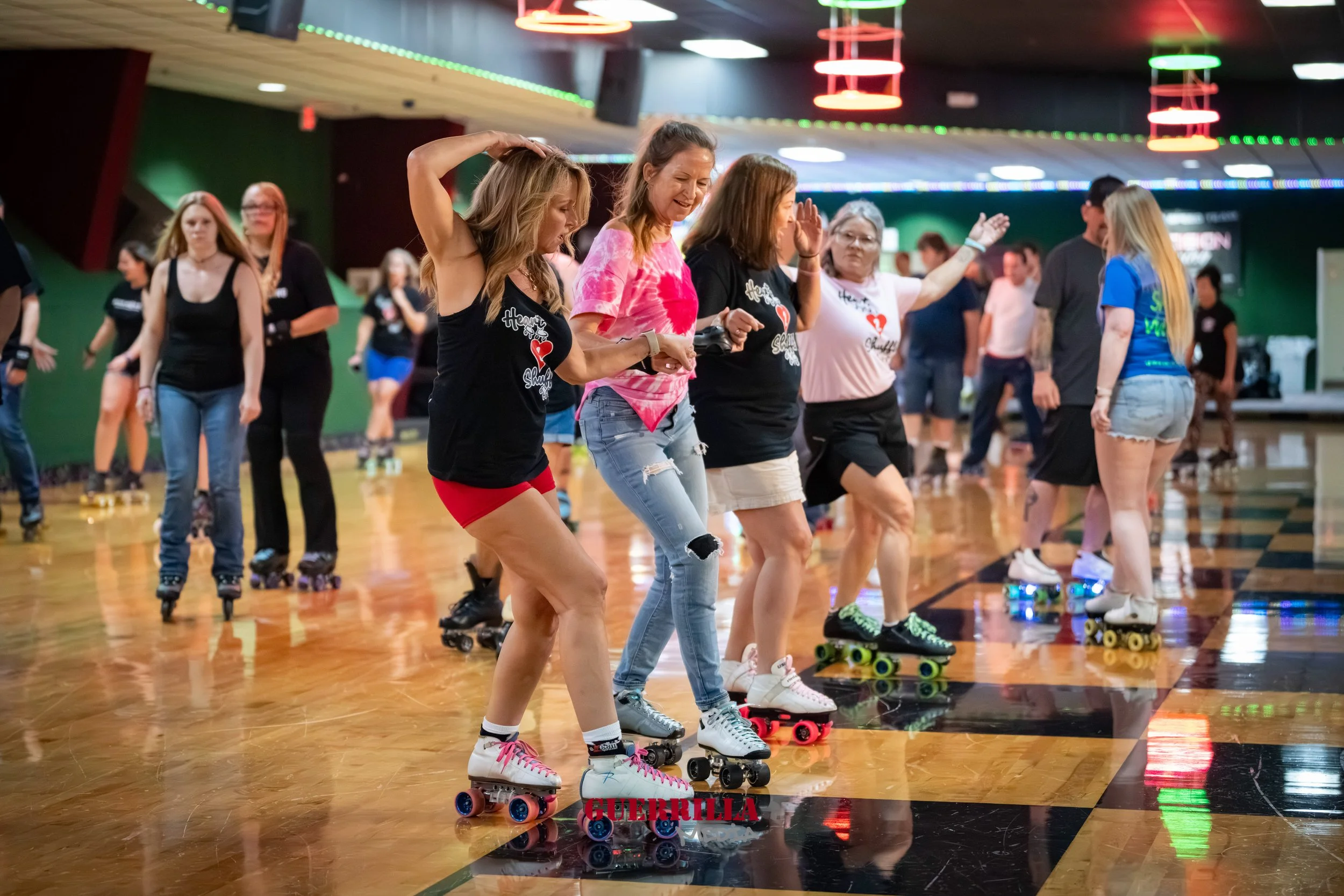 Group of women roller skating indoors, some smiling and holding hands, with colorful lights overhead and a polished wooden floor.