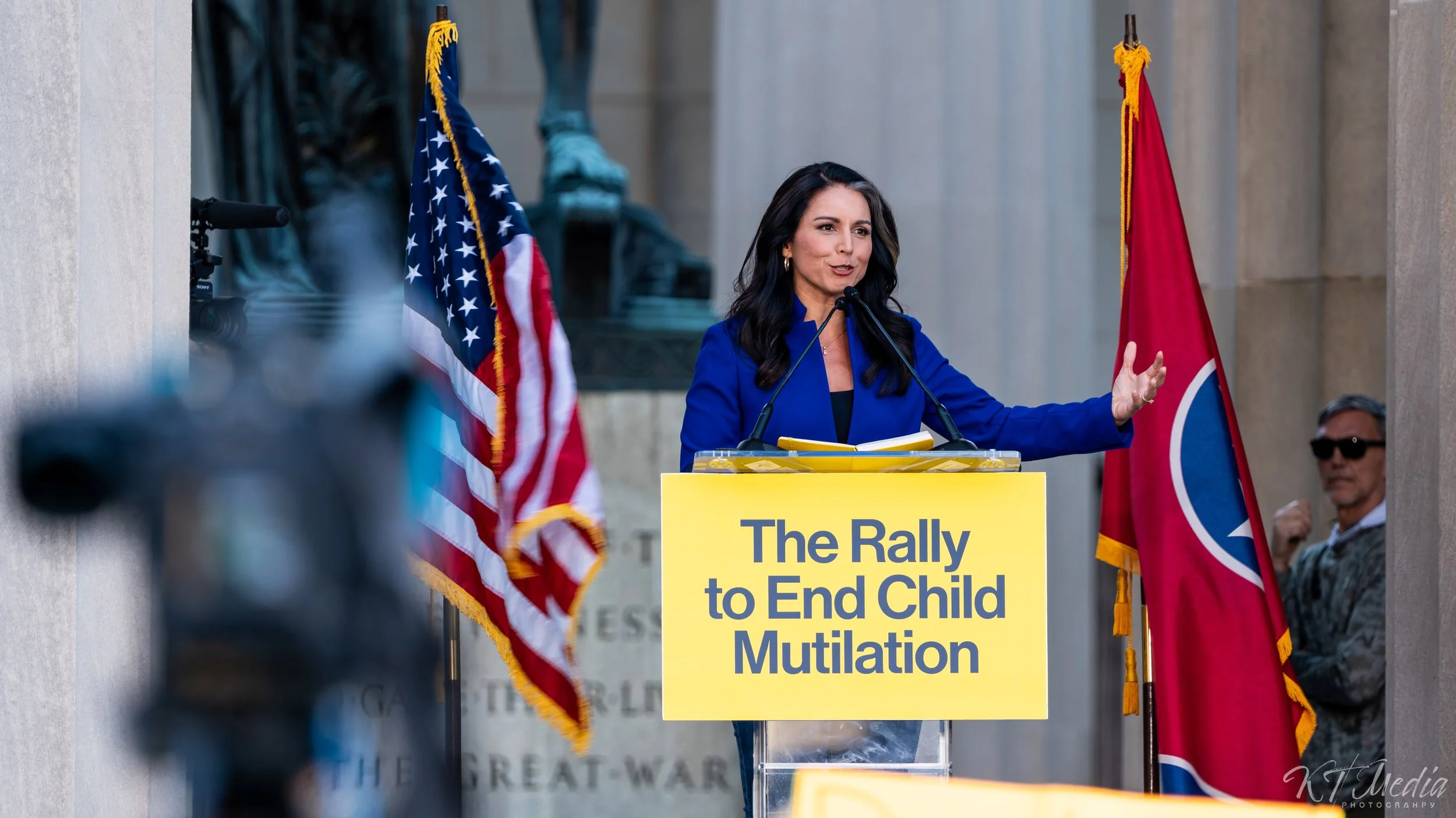 A woman speaking at a rally with a sign that reads 'The Rally to End Child Mutilation'; American flags are placed on either side of the podium.