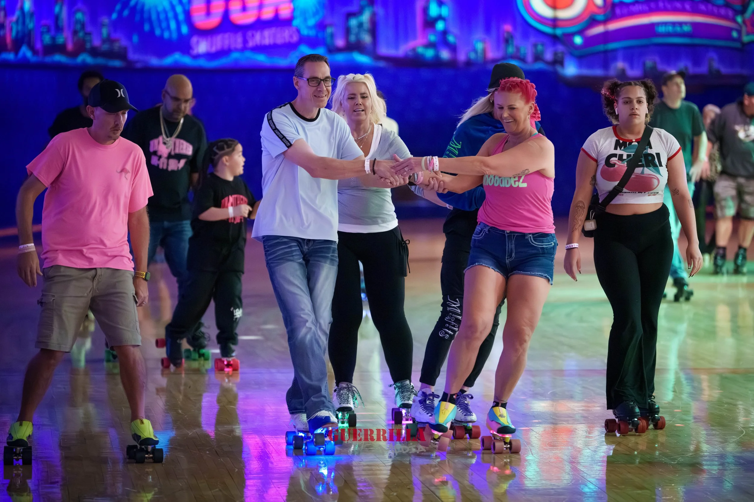 People roller skating in a brightly lit indoor skating rink, with colorful neon lights and a digital display in the background.