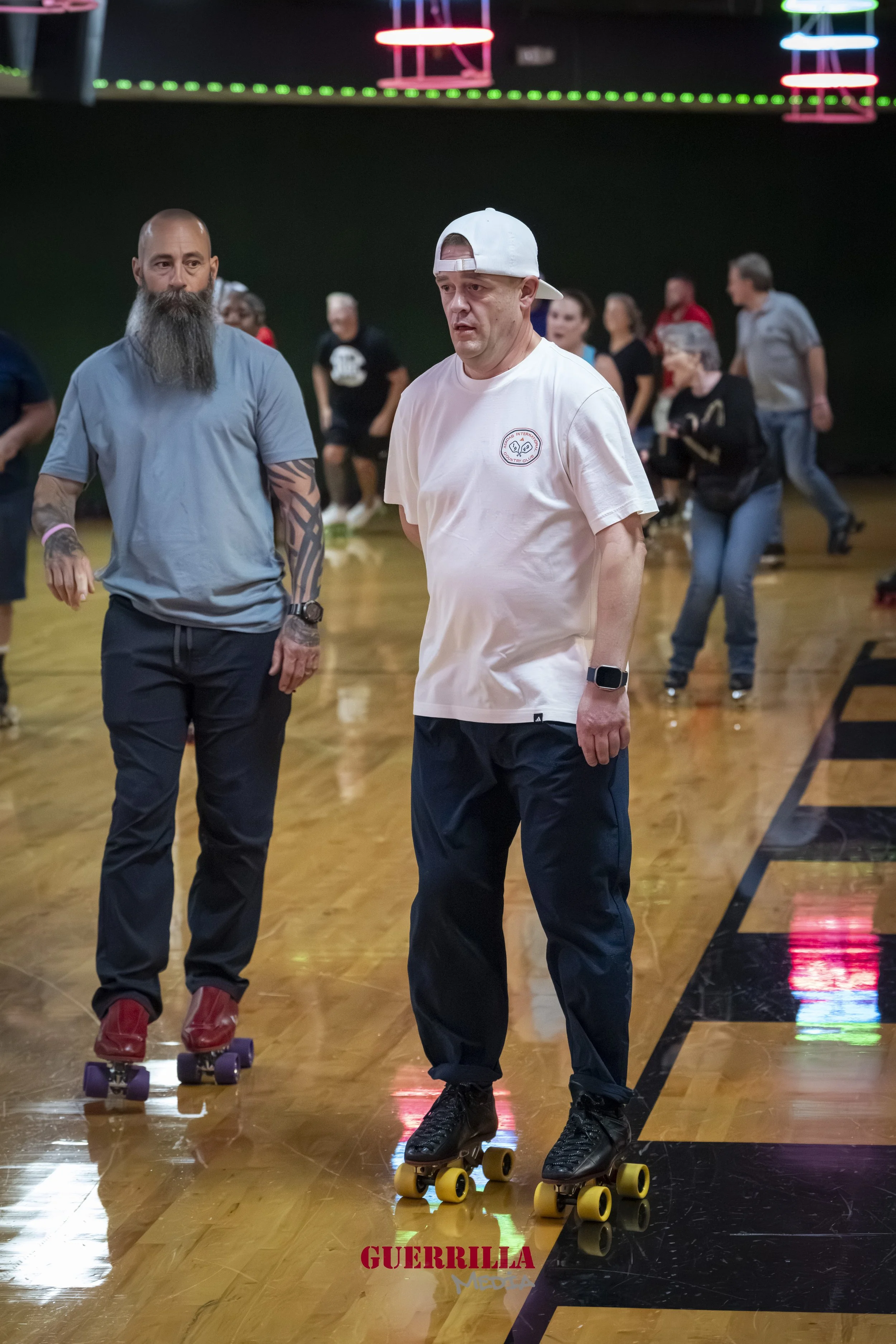 Two men roller skating in an indoor rink, with people in the background. One man has a beard and tattoos, and is wearing a blue shirt and dark pants. The other man is wearing a white t-shirt, a white cap worn backwards, and dark pants, with a smartwa