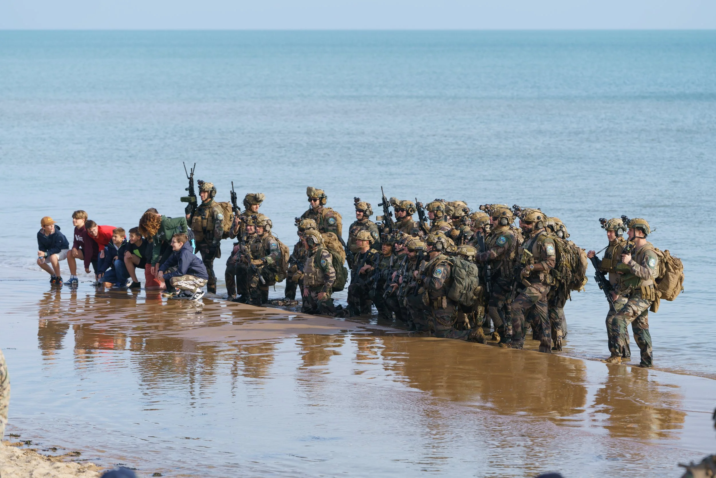 Military personnel in full gear standing knee-deep in water, kneeling and crouching in formation on a beach, with children nearby, during daytime.