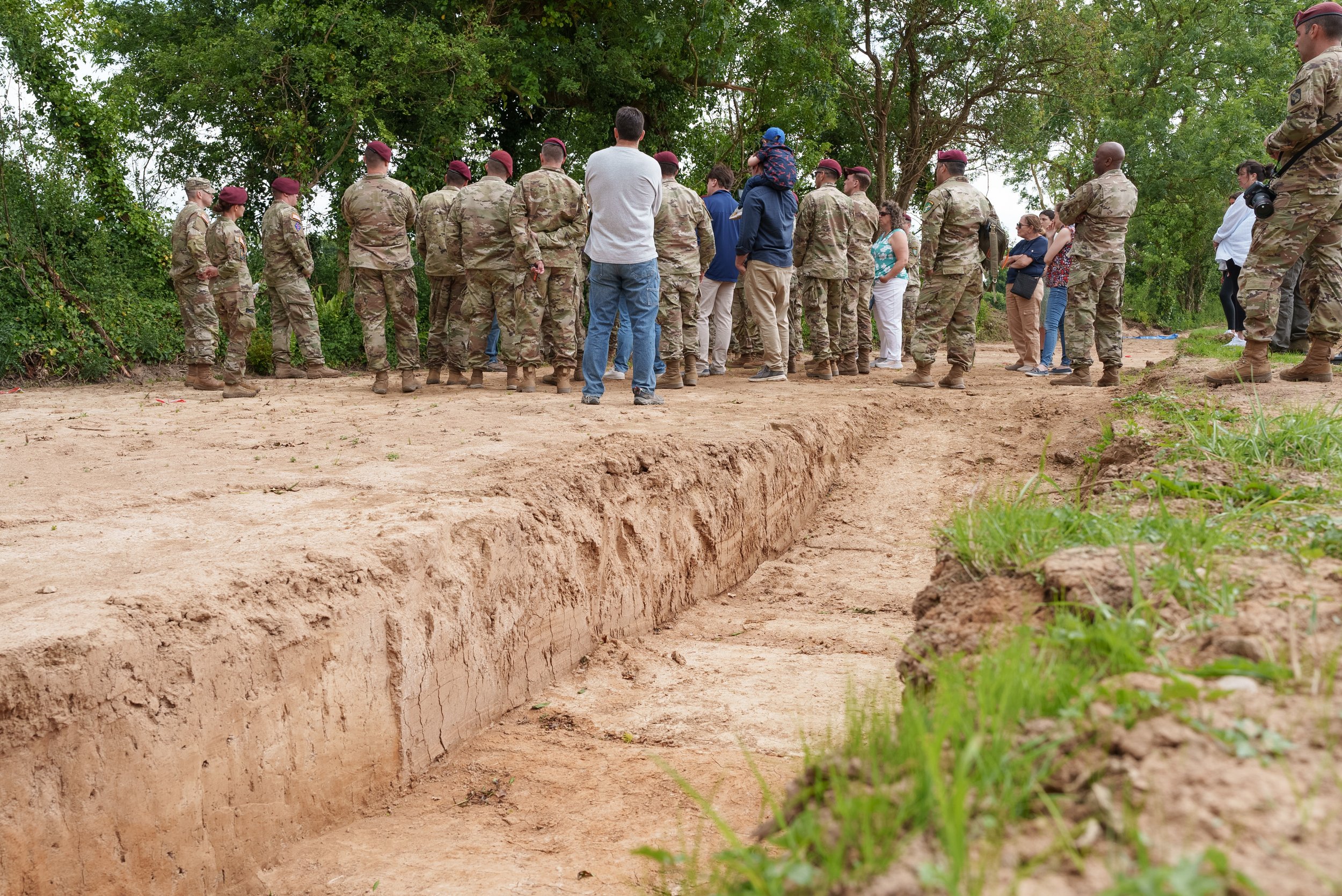 A group of soldiers and civilians standing around a trench or excavated area outdoors during the day, with green trees in the background.