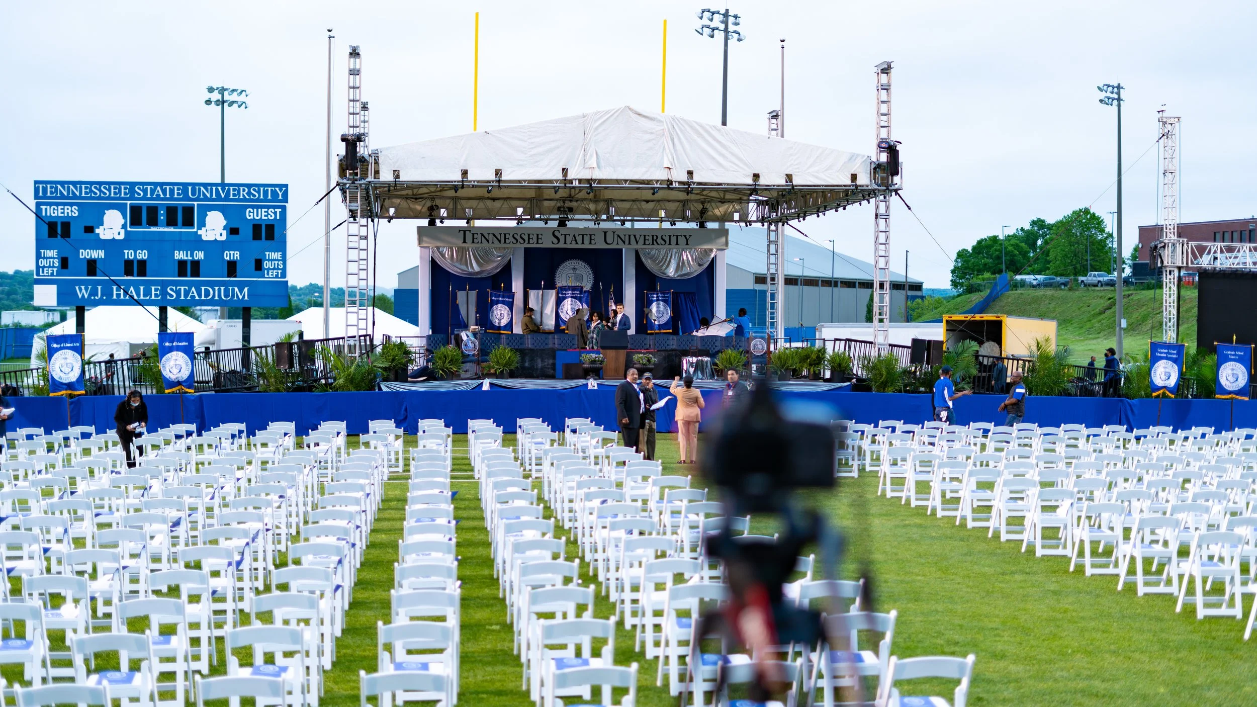 An outdoor graduation ceremony setup at Tennessee State University on a grass field, with white chairs arranged in rows, a stage with a canopy, and a large electronic scoreboard displaying the university's name and event details.