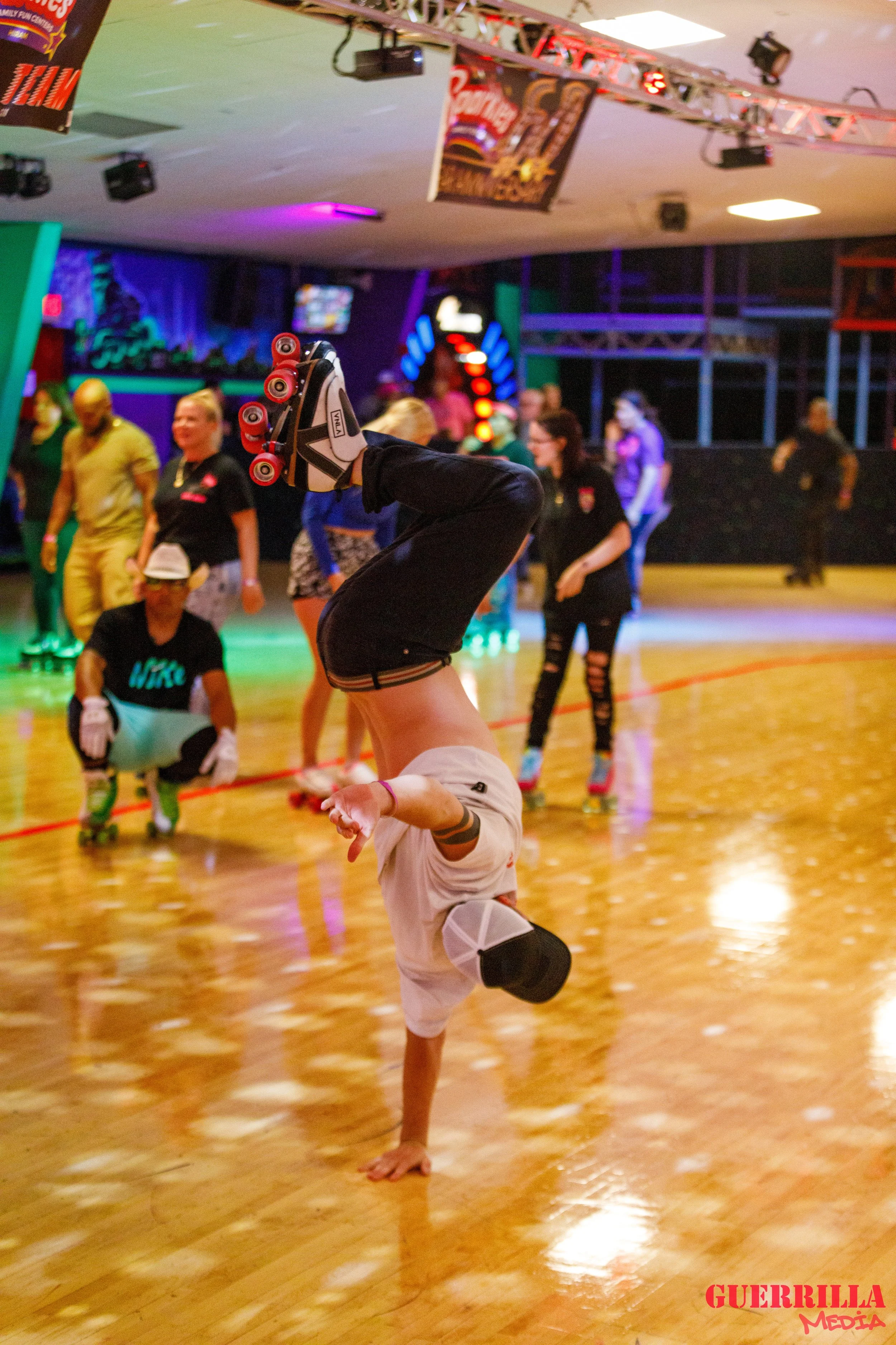 A person performing a handstand on roller skates in an indoor roller skating rink with colorful lights and other skaters in the background.