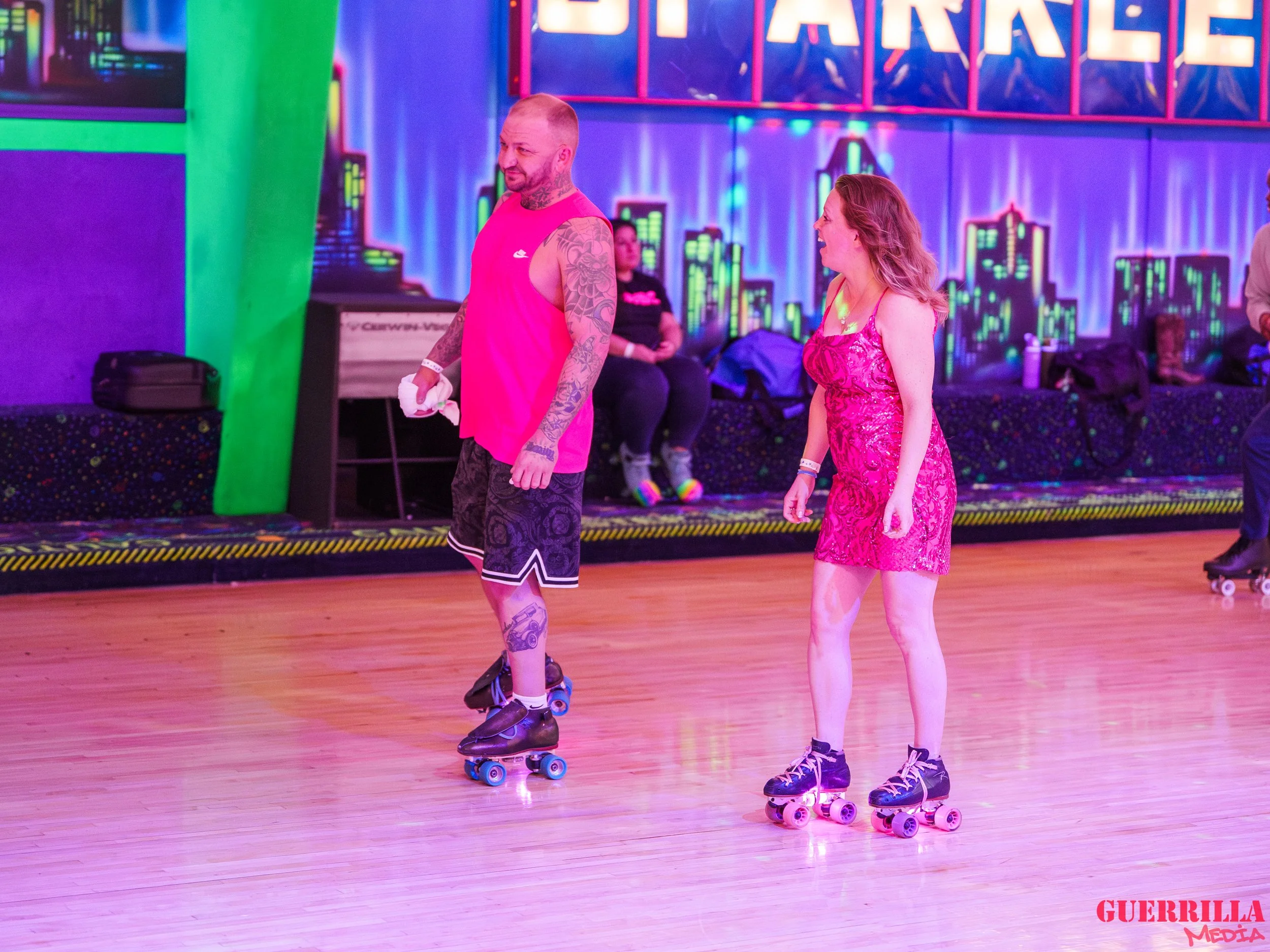 Two people roller skating in an indoor rink with neon lights and a cityscape mural in the background. One man with tattoos is wearing a pink sleeveless shirt and black shorts, and a woman in a pink dress is skating nearby.