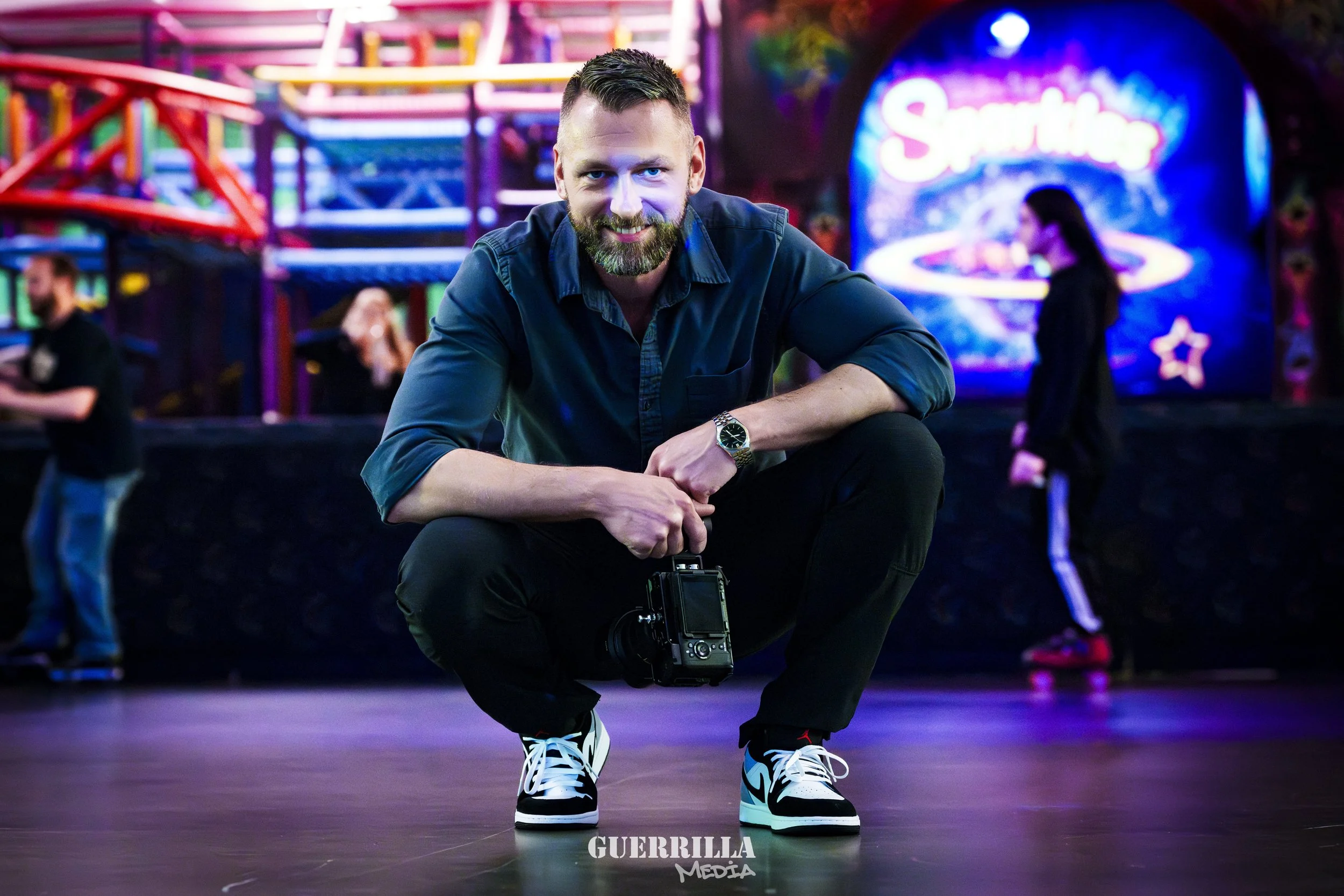 Man in a dark shirt and sneakers squatting on a roller rink floor, holding a camera, with a colorful amusement park ride and a neon sign in the background.