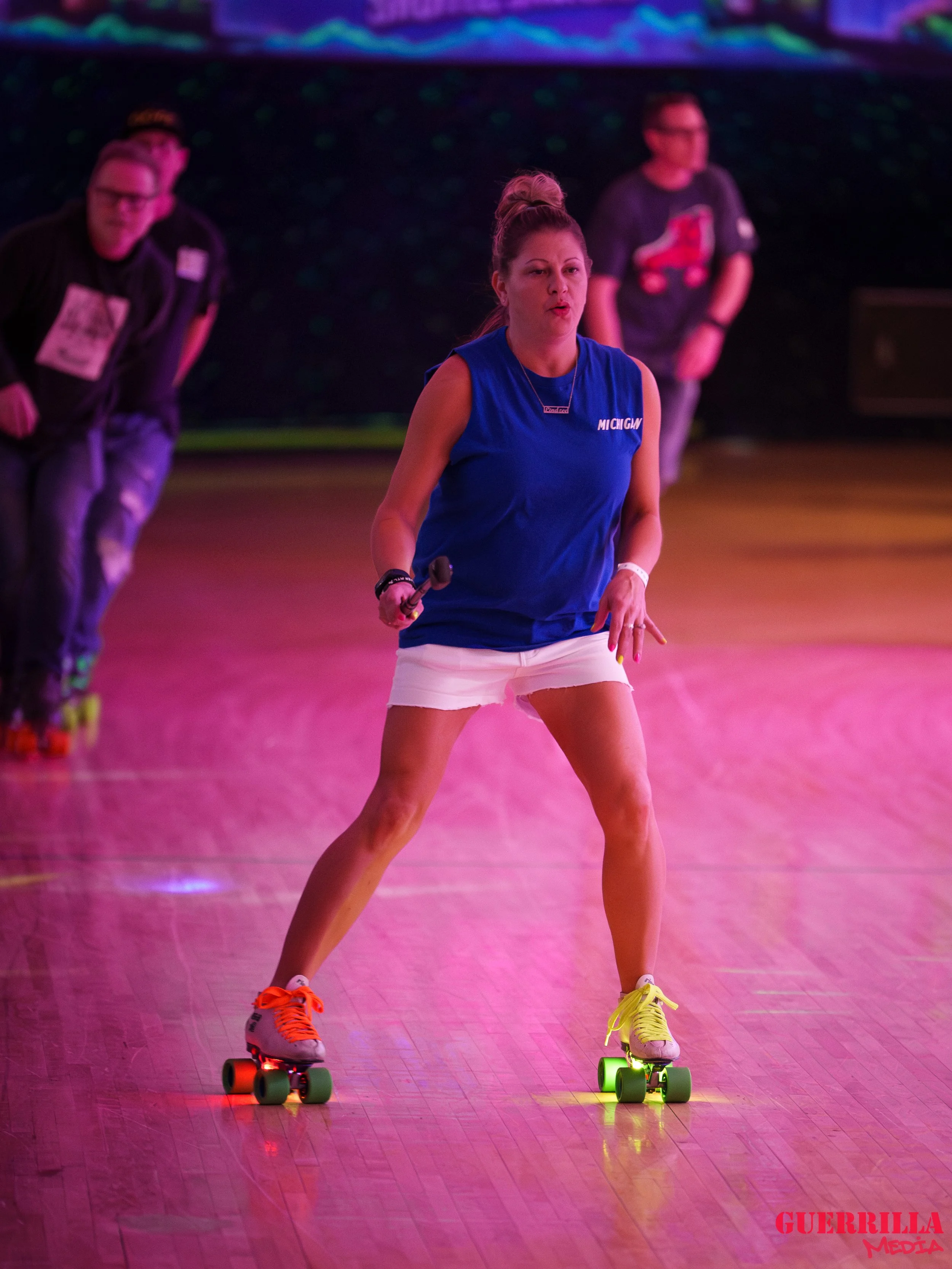 A woman roller skating indoors with a neon-lit background, wearing a Michigan sleeveless shirt, white shorts, and colorful roller skates, holding a microphone.