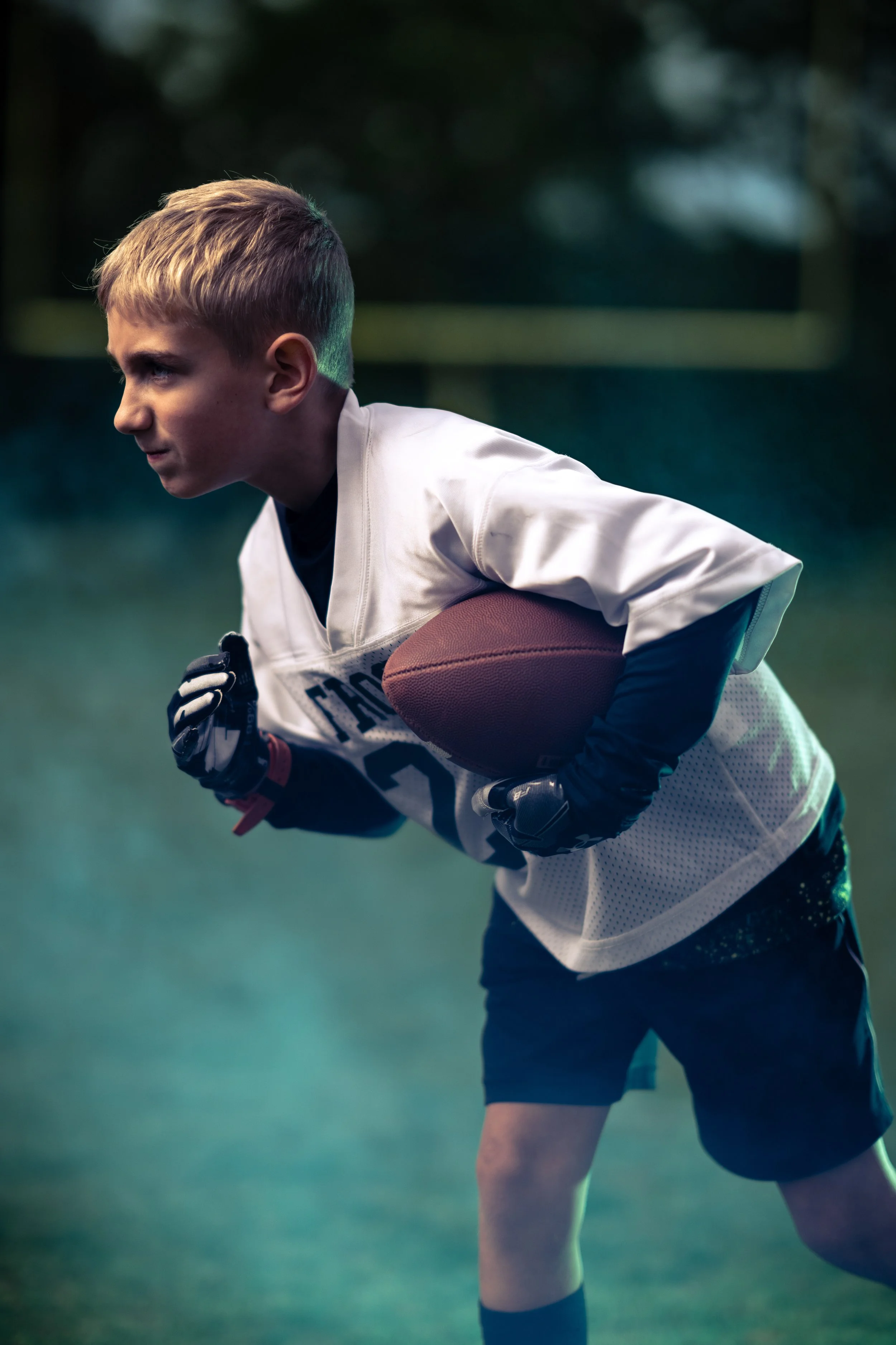 Young boy in football gear holding a football and running on a football field.