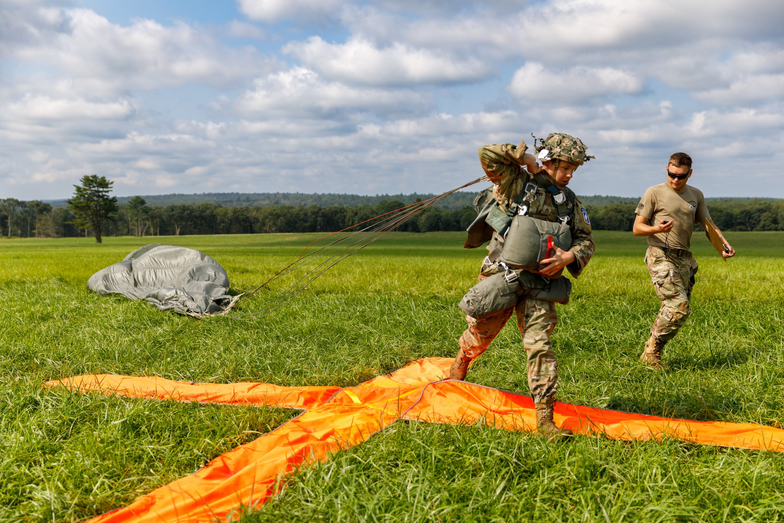 Military personnel preparing a parachute on a grassy field with a cloudy sky in the background.