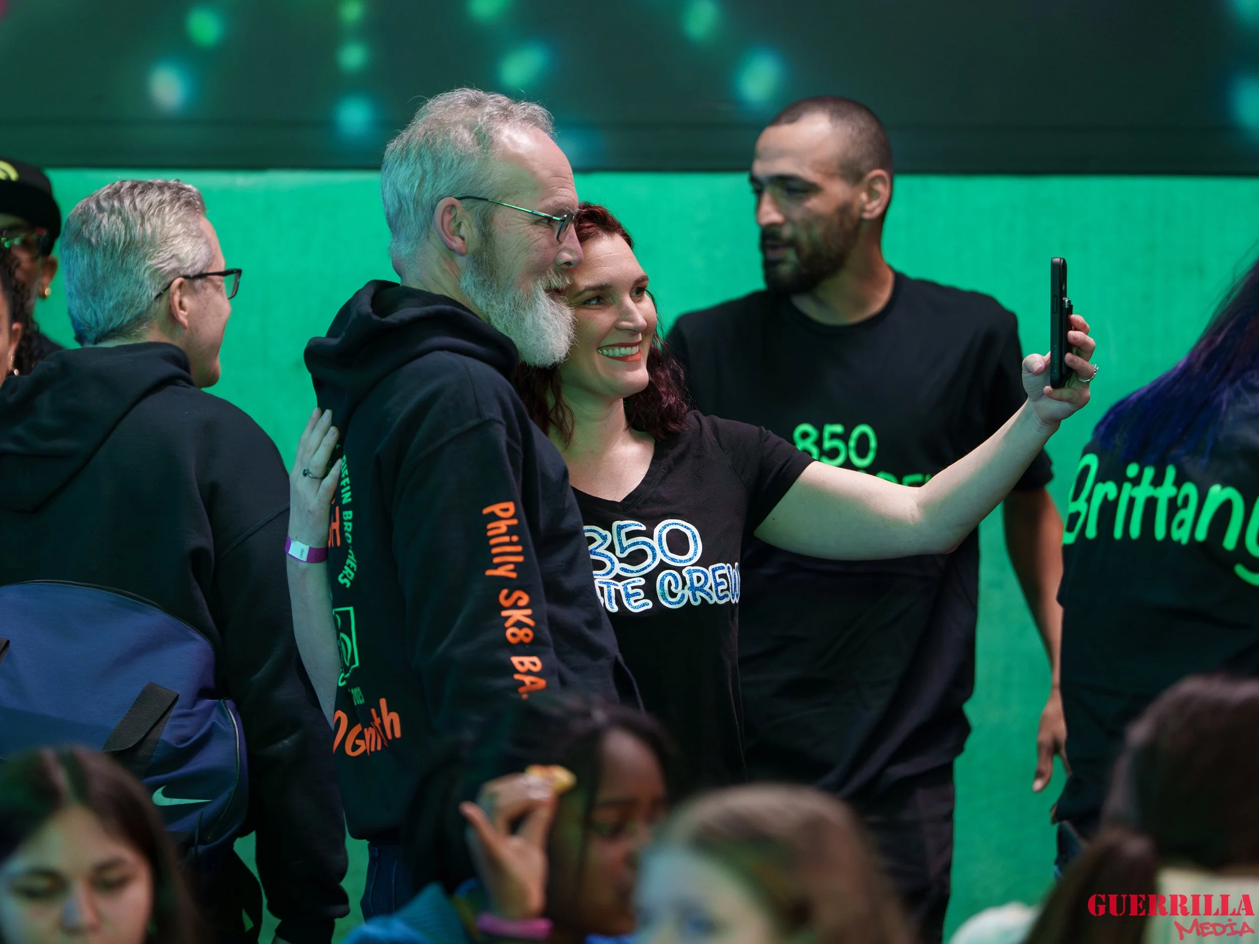 A group of people taking a selfie indoors with a green background, some wearing black T-shirts with white and green text, and smiling.