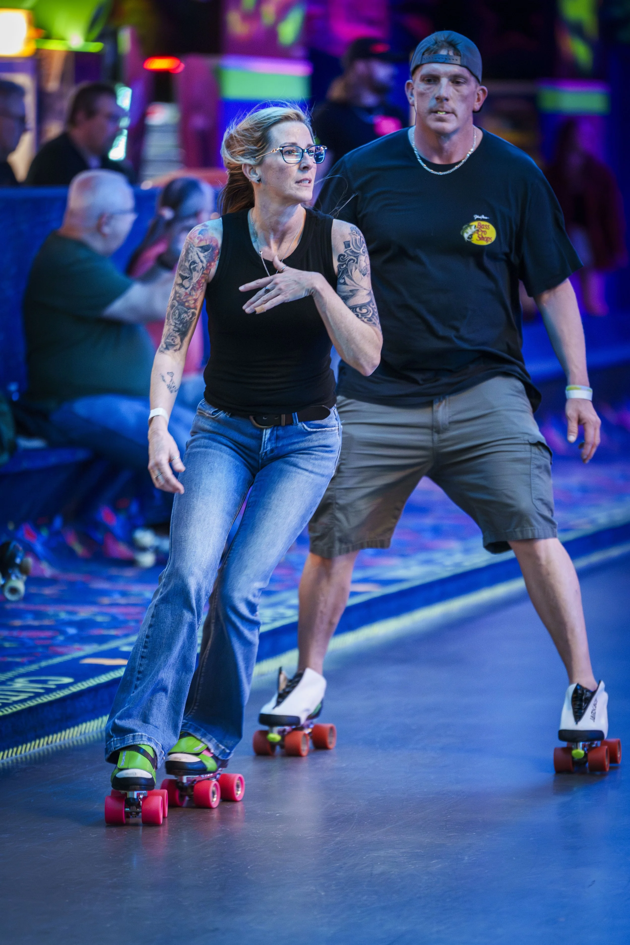 A woman with tattoos and glasses skating indoors on roller skates, with a man in a baseball cap and shorts skating behind her, at an environment with colorful neon lights.