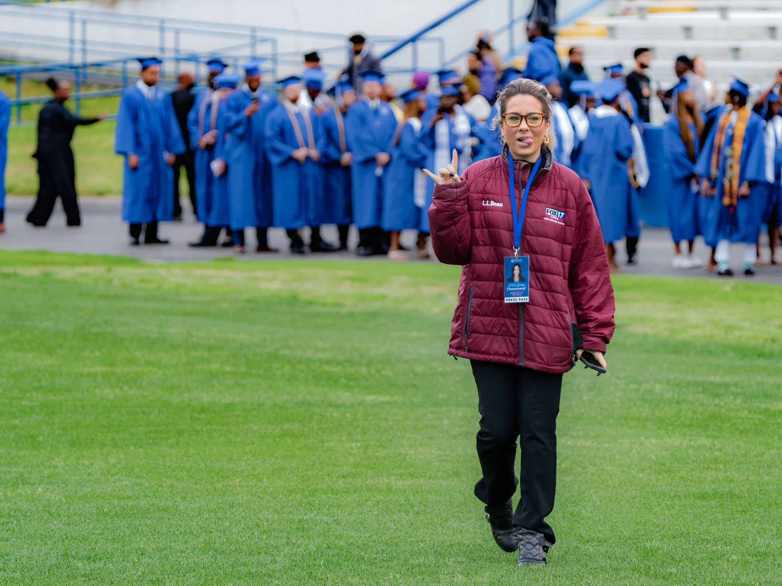 A woman in a maroon jacket with around her neck, an ID badge, and holding a phone, walking on grass in front of a group of people in blue graduation gowns and caps gathered at a stadium.