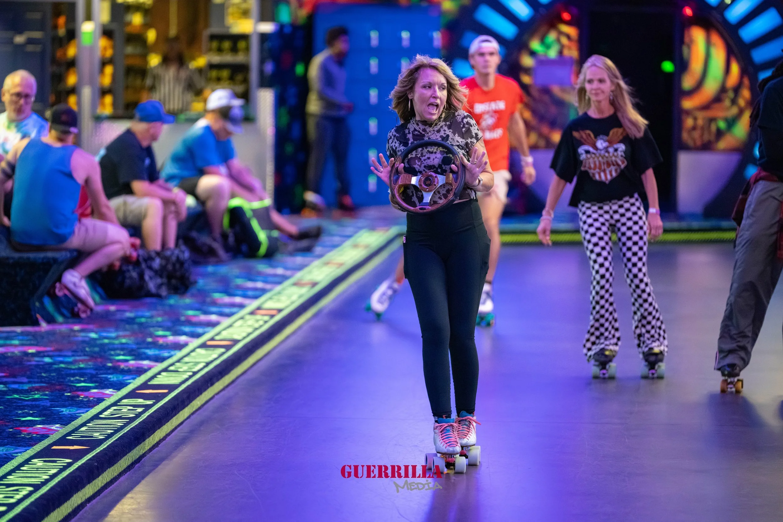 Woman roller skating on an indoor roller rink, holding a steering wheel prop, with other skaters and spectators in the background.