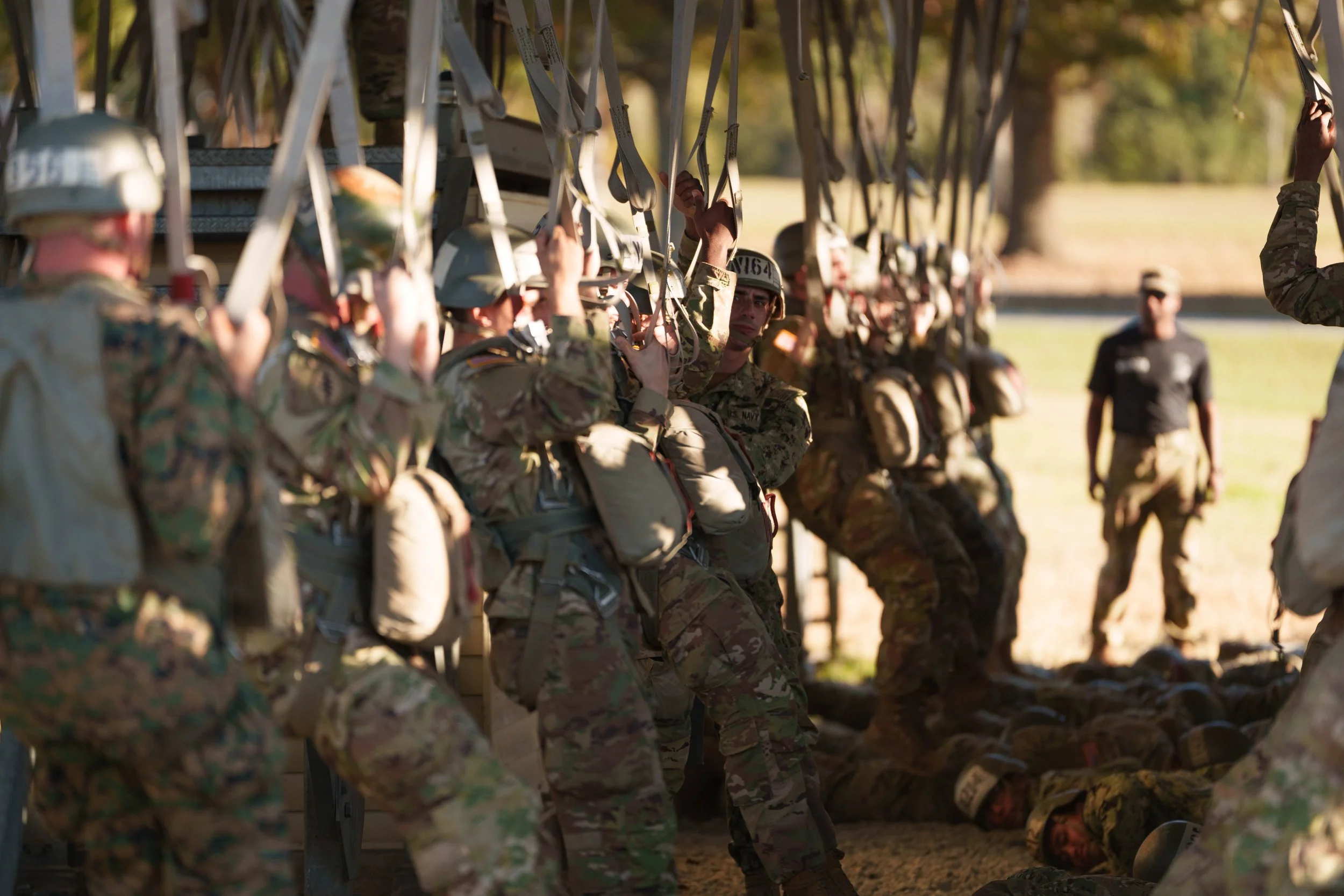 Military personnel in camouflage uniforms and helmets prepare to deploy from an aircraft, with some soldiers standing and others lying on the ground under the aircraft.