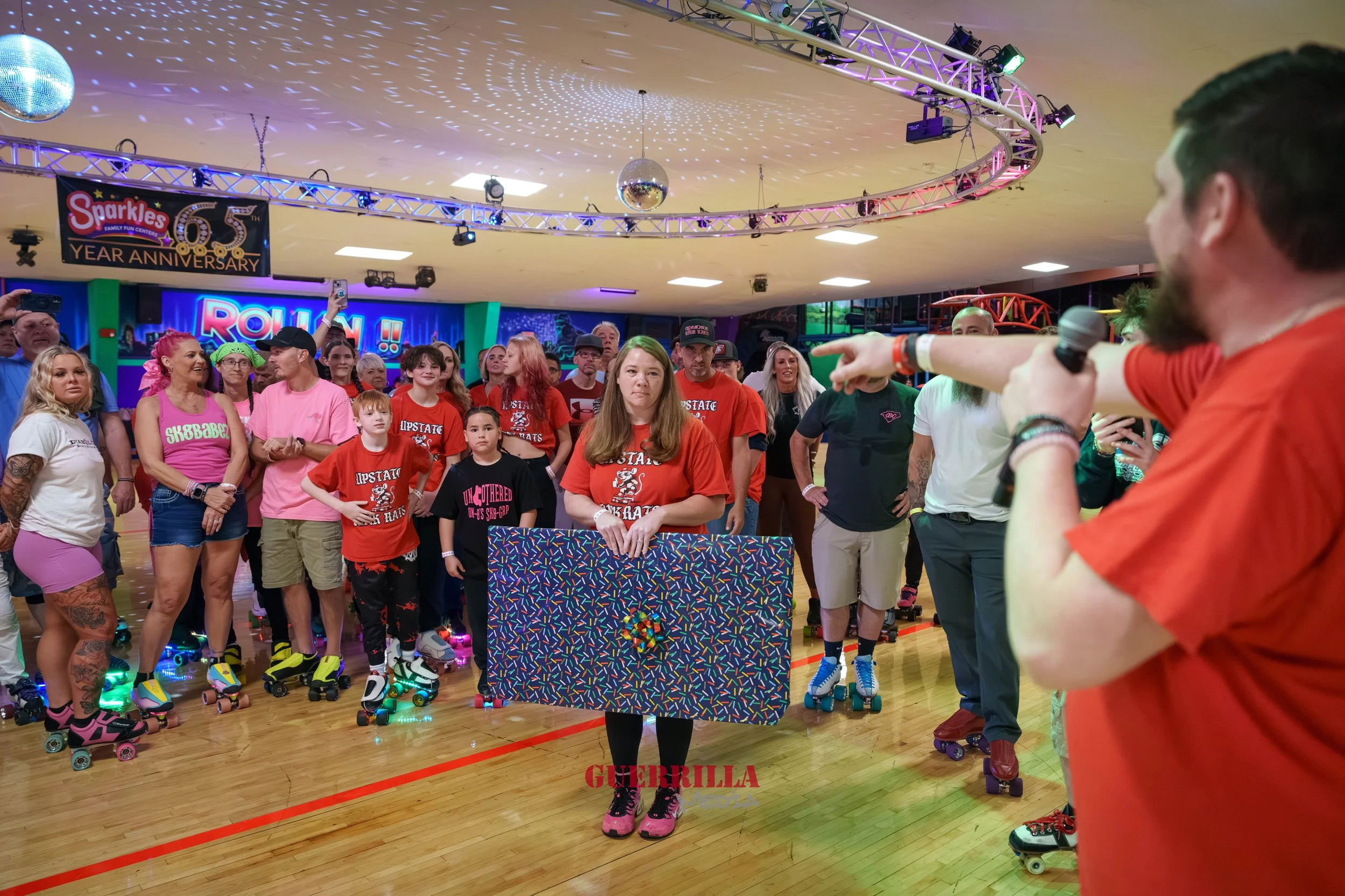 A group of people on roller skates gathered in an indoor roller skating rink, with a woman in the center holding a large, colorful gift box. A man with a microphone appears to be addressing the crowd, pointing towards the woman. The background featur