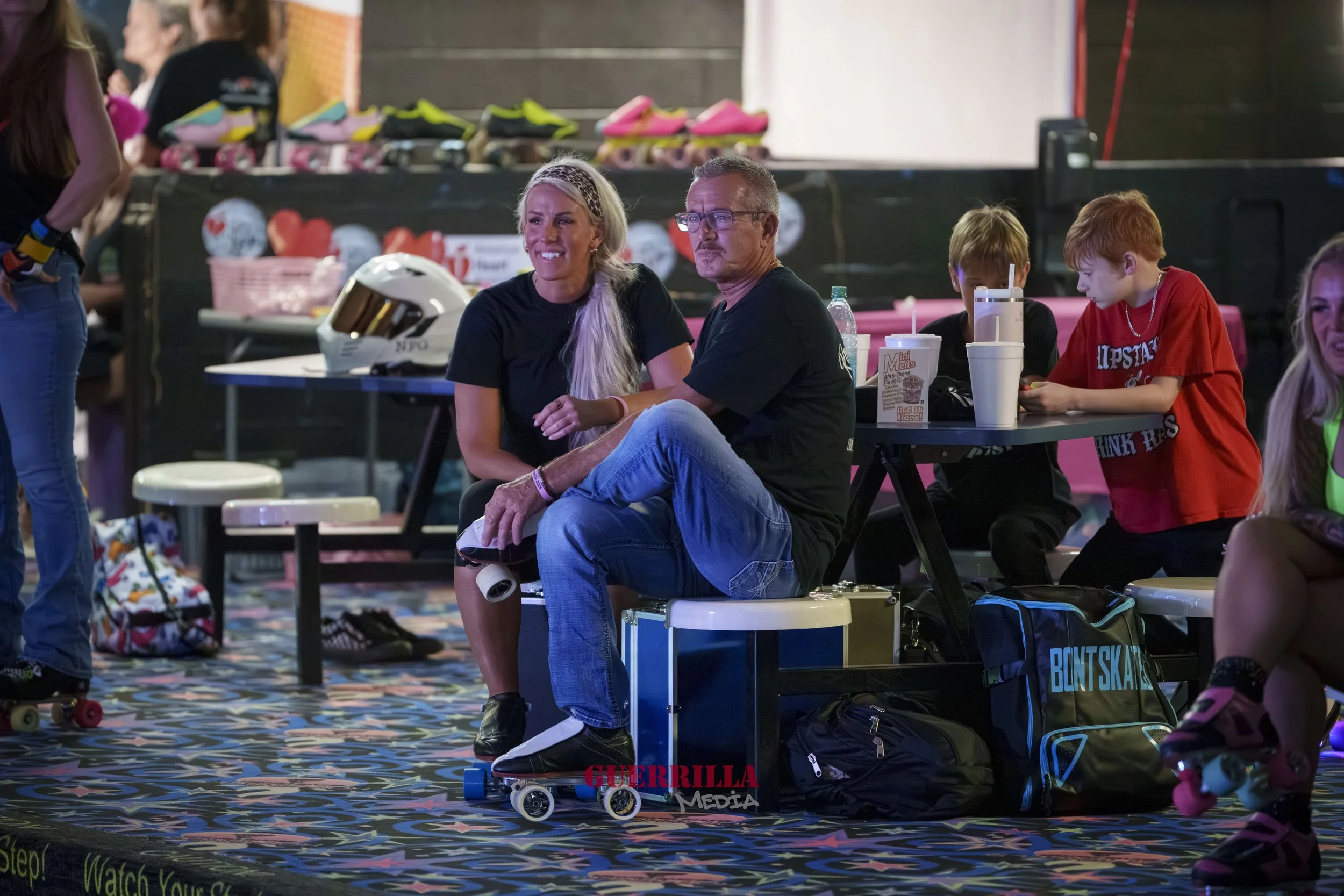 People at an indoor roller skating rink. Two adults sit on benches, one woman with long blonde hair and one man with glasses, with children sitting nearby. Skates and helmets are visible, along with skating gear and a patterned carpet.