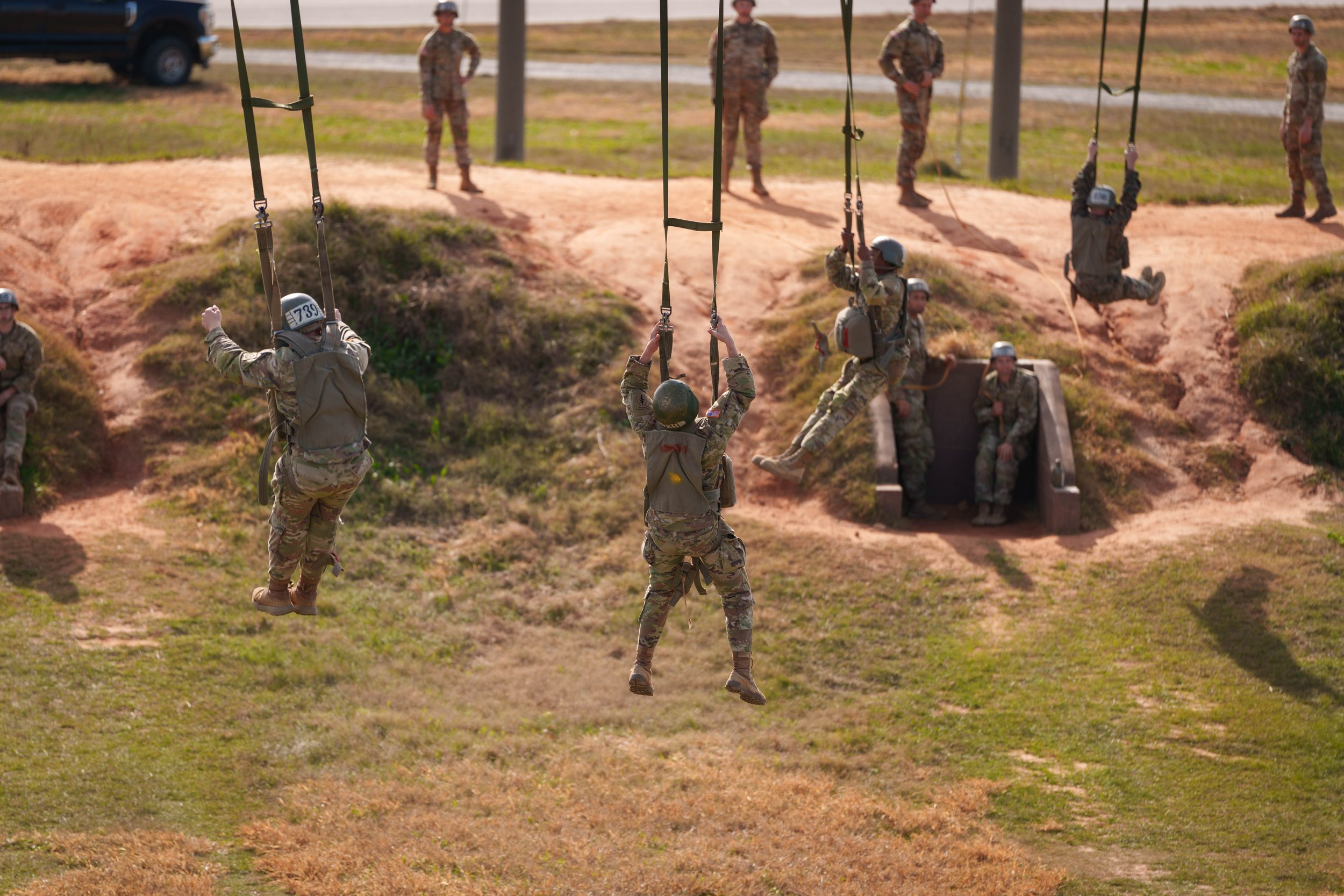 Military personnel participating in a training exercise, swinging on ropes and preparing to land on the ground with others watching.
