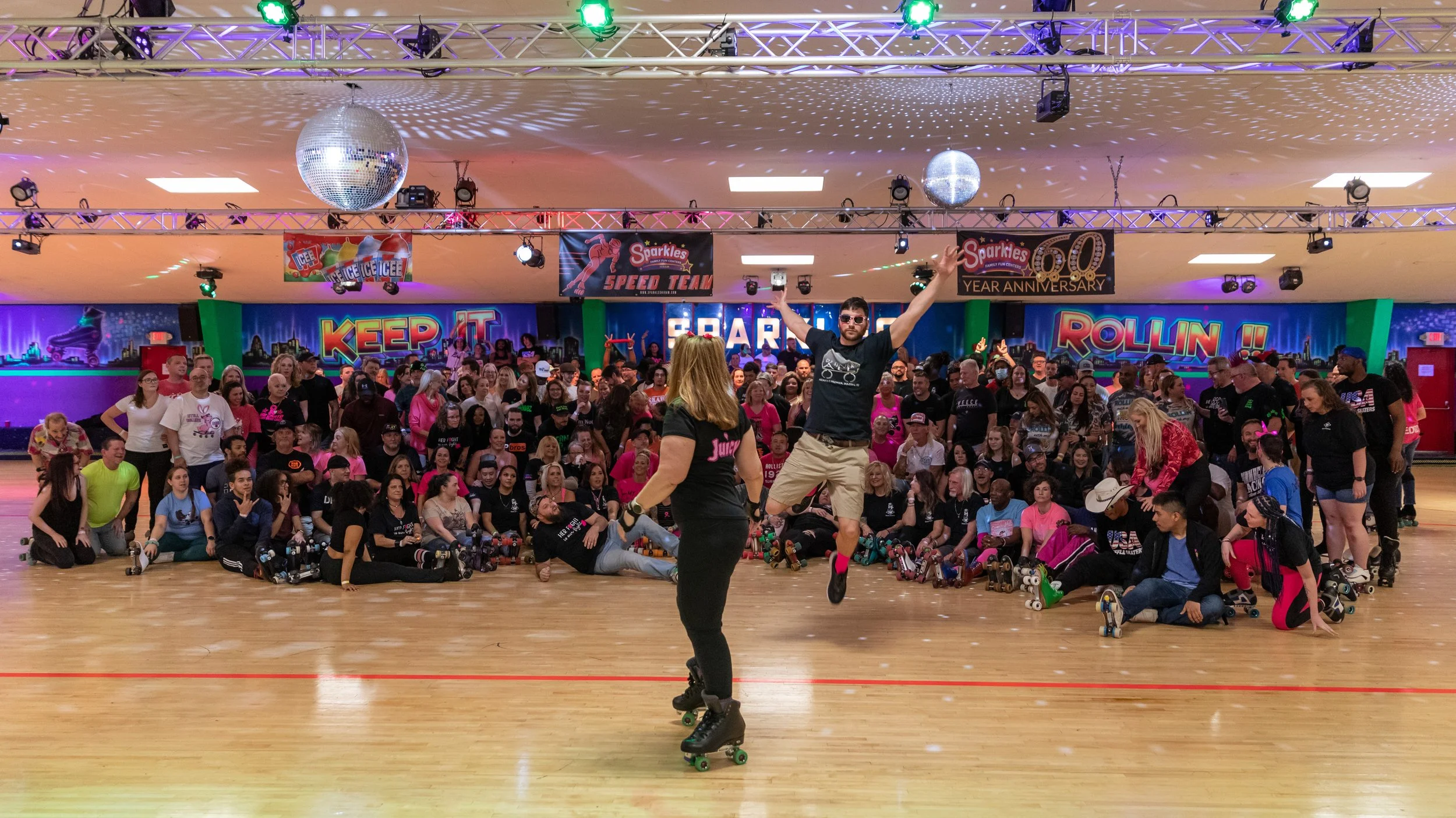 A large group of people gathered in a roller skating rink, with two roller skaters jumping in the foreground. The venue is decorated with colorful signs and banners, including one celebrating a 50-year anniversary and another with a theme of keeping 