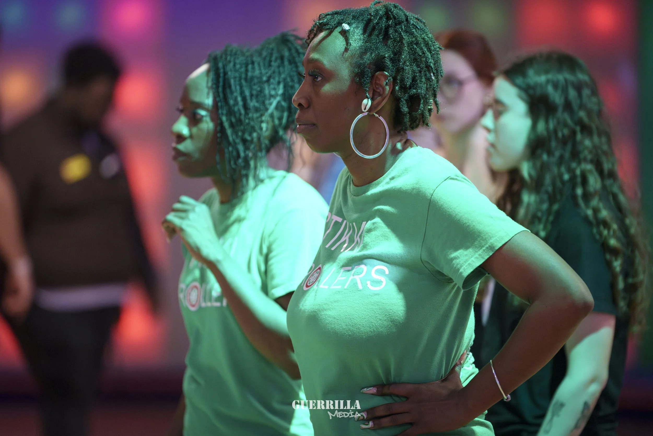 Group of women standing together in a dimly lit room with colorful lighting, some with their eyes closed or looking serious, wearing casual T-shirts and large hoop earrings.