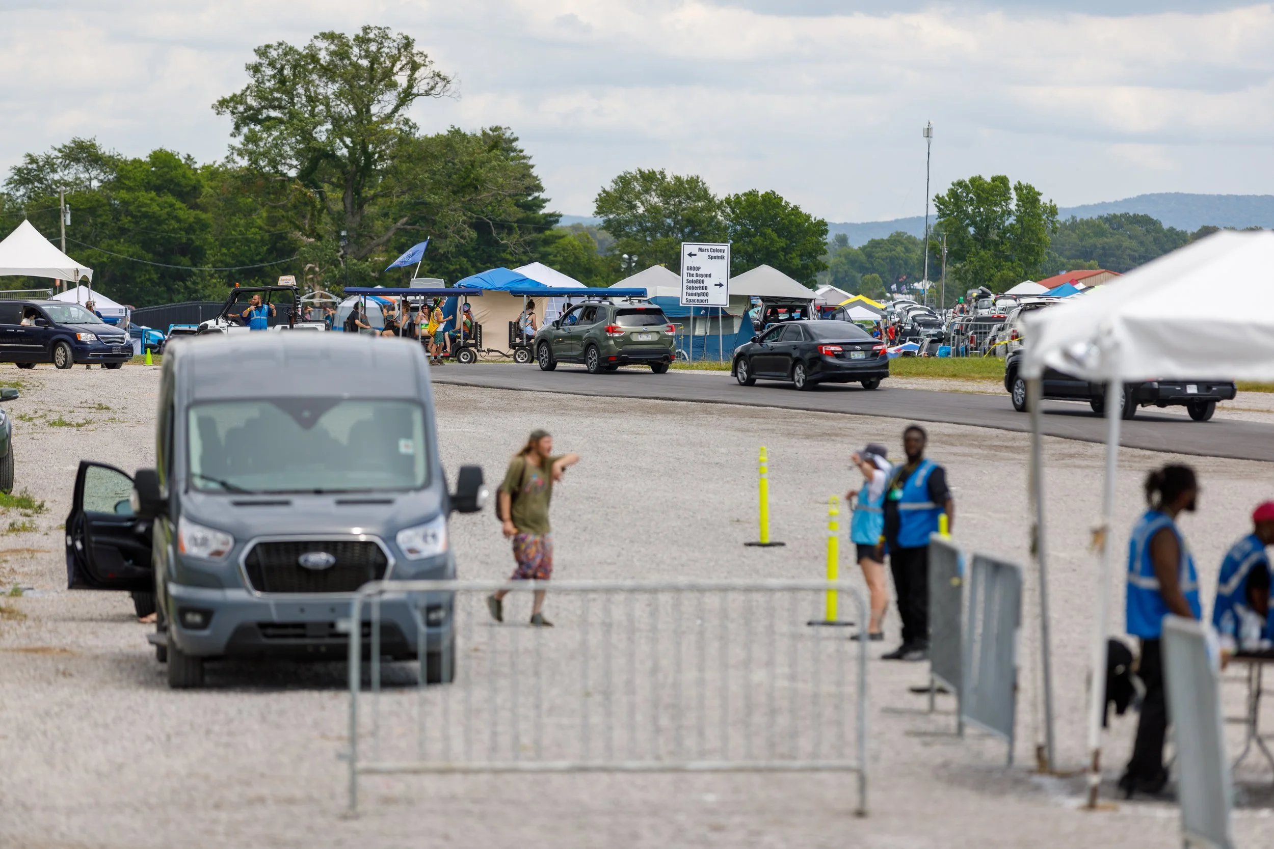 Outdoor scene with tents, cars, and people, some wearing vests, in a parking area or event space, with trees and cloudy sky in the background