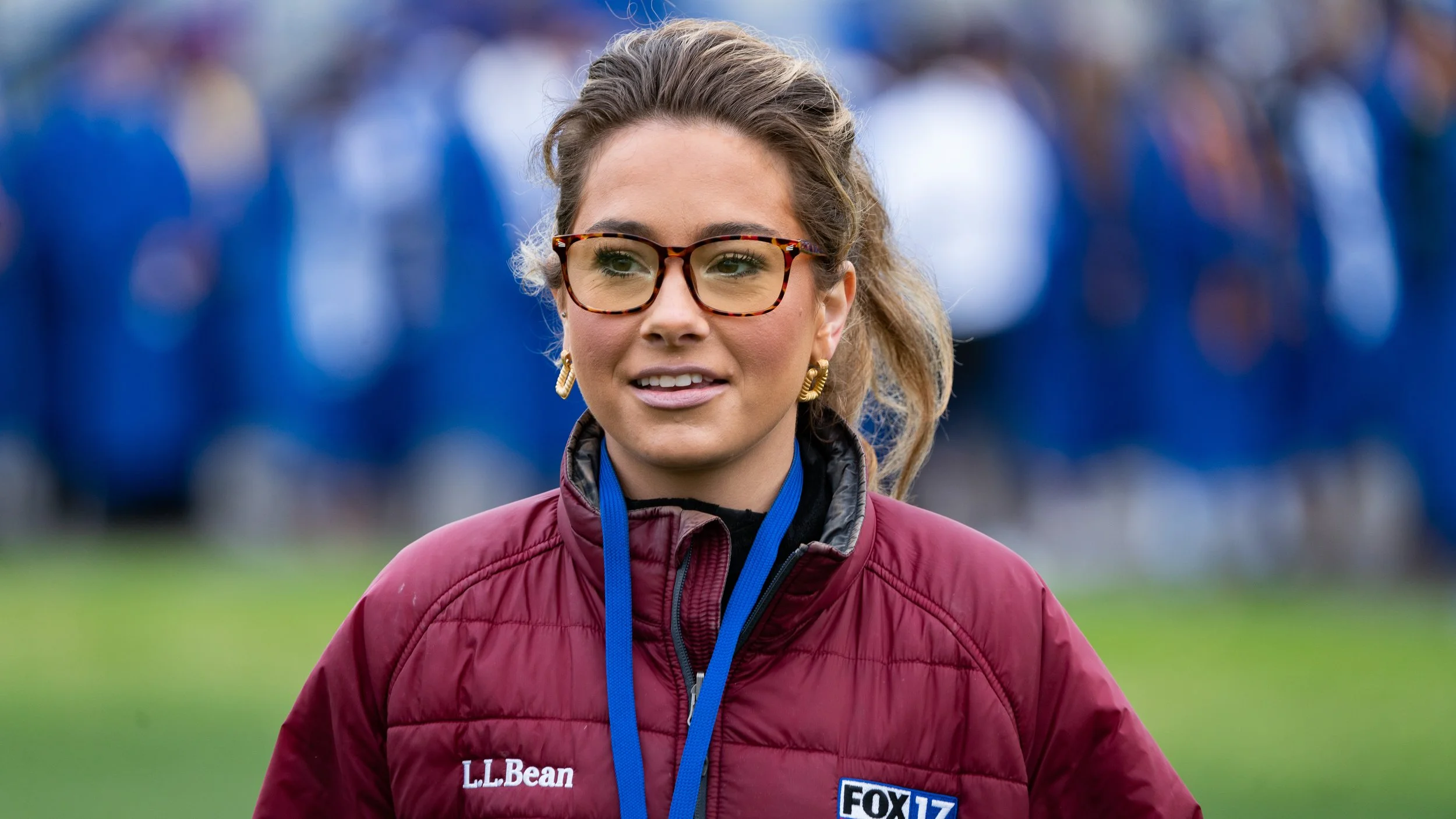 A woman wearing glasses, earrings, and a red jacket with a 'L.L. Bean' logo and a 'FOX 17' patch. She has a lanyard around her neck and is outdoors, with a blurred crowd in the background.