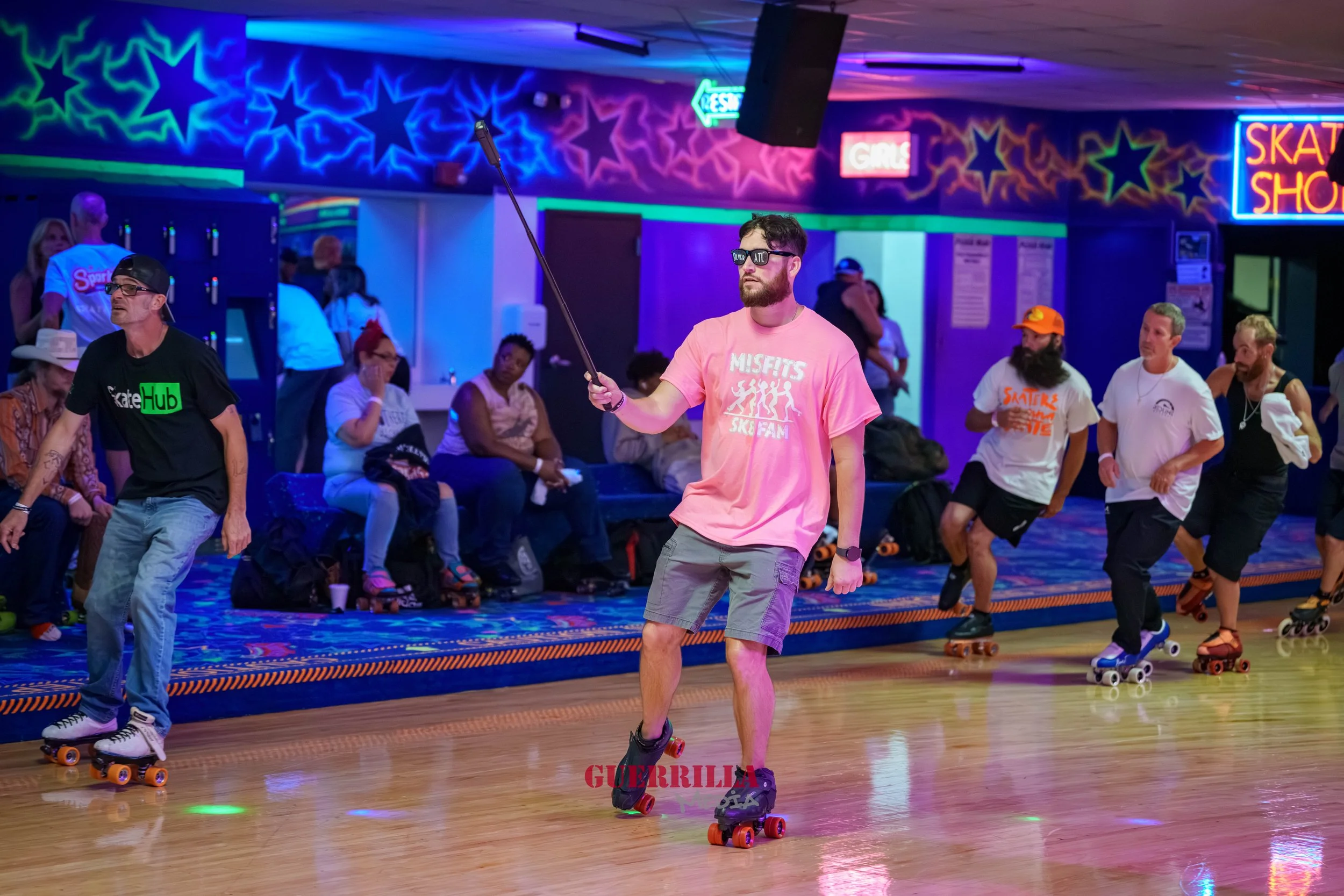 A man wearing sunglasses, a pink Misfits t-shirt, and gray shorts roller skating at an indoor skating rink with neon lights. He is holding a selfie stick. In the background, other skaters and seated spectators are visible, with neon signs and star-sh