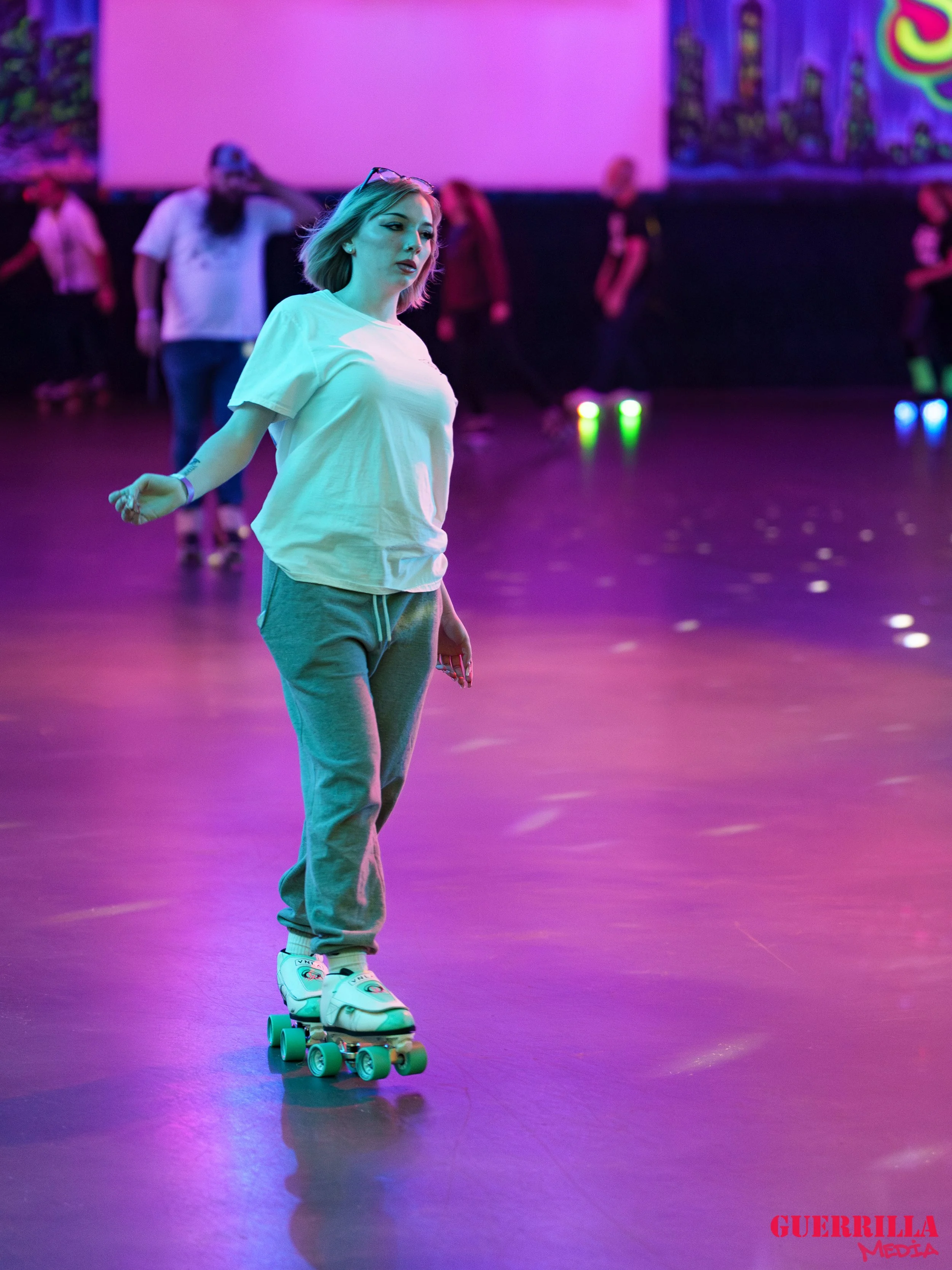 A young woman roller skating in a dimly lit roller rink with colorful neon lights, wearing a white t-shirt, gray sweatpants, and white roller skates.