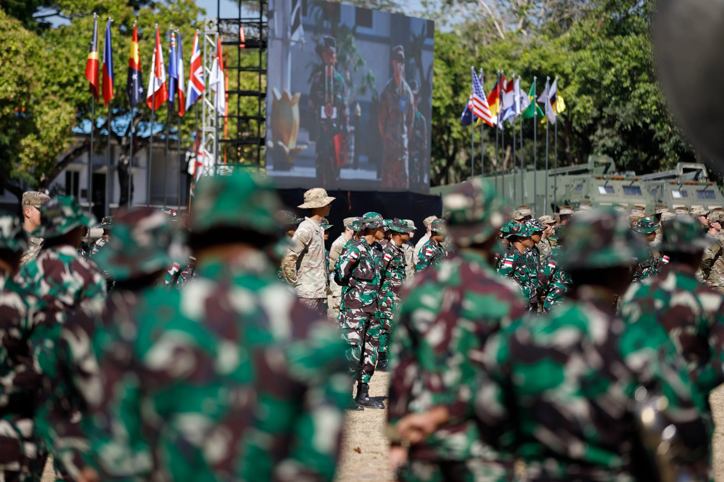 Military personnel gathered outdoors for a ceremony or event, with flags displayed in the background and a large screen showing participants.