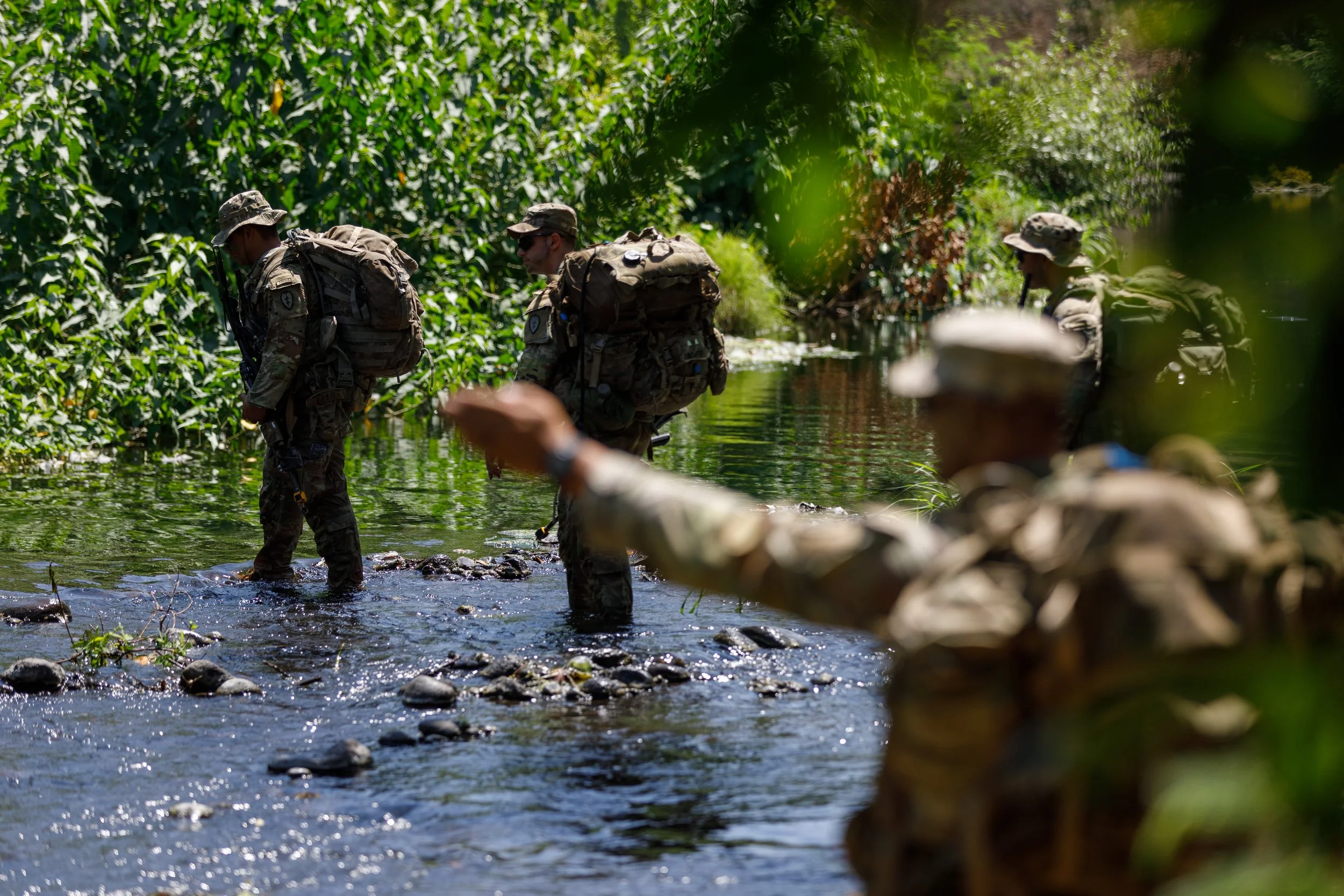 Military personnel wading through a shallow river in a lush green forest, wearing camouflage uniforms and backpacks.