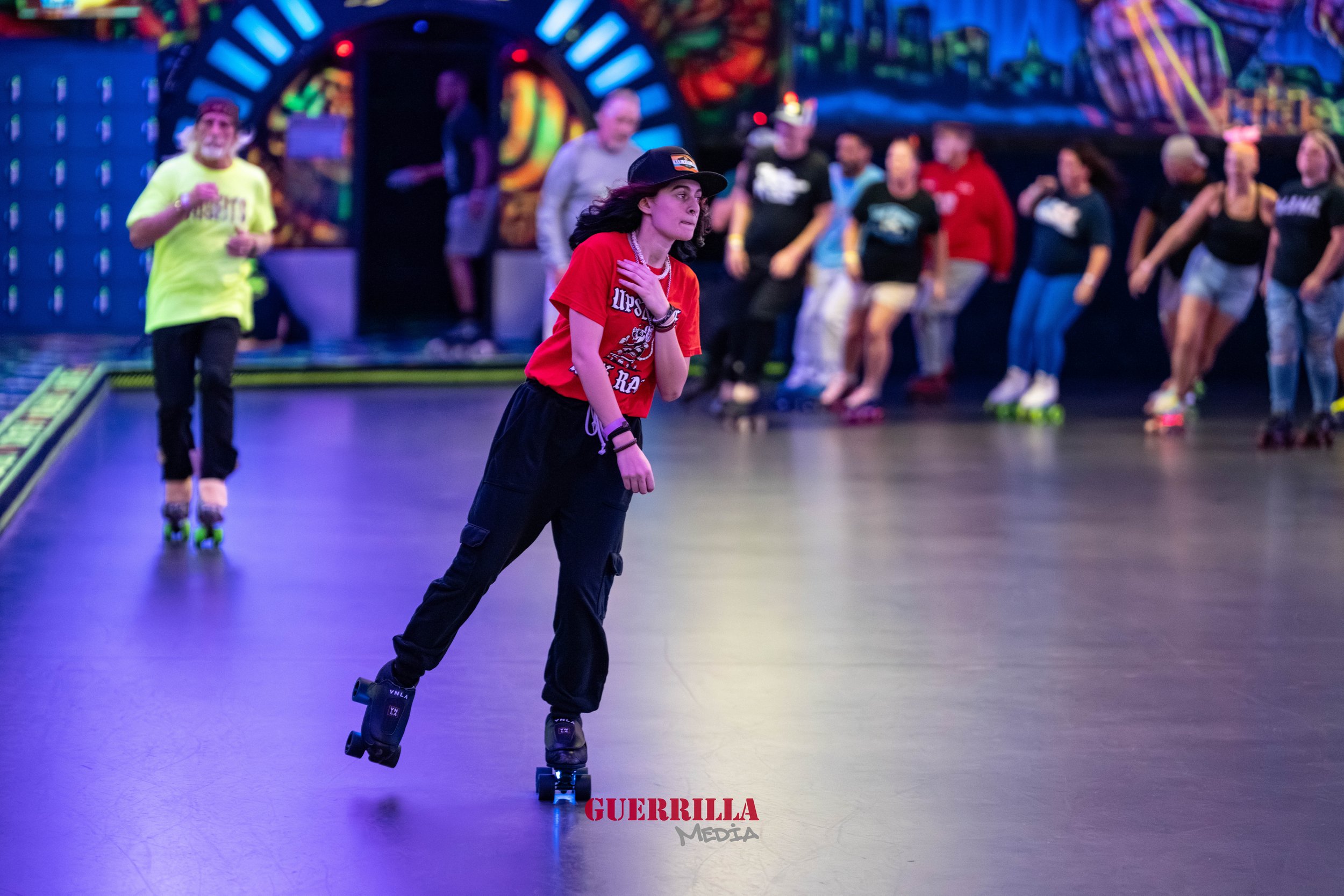 A young woman wearing a red shirt and black pants roller skating at an indoor roller rink, with a crowd of people in the background. The scene is illuminated with colorful neon lights.