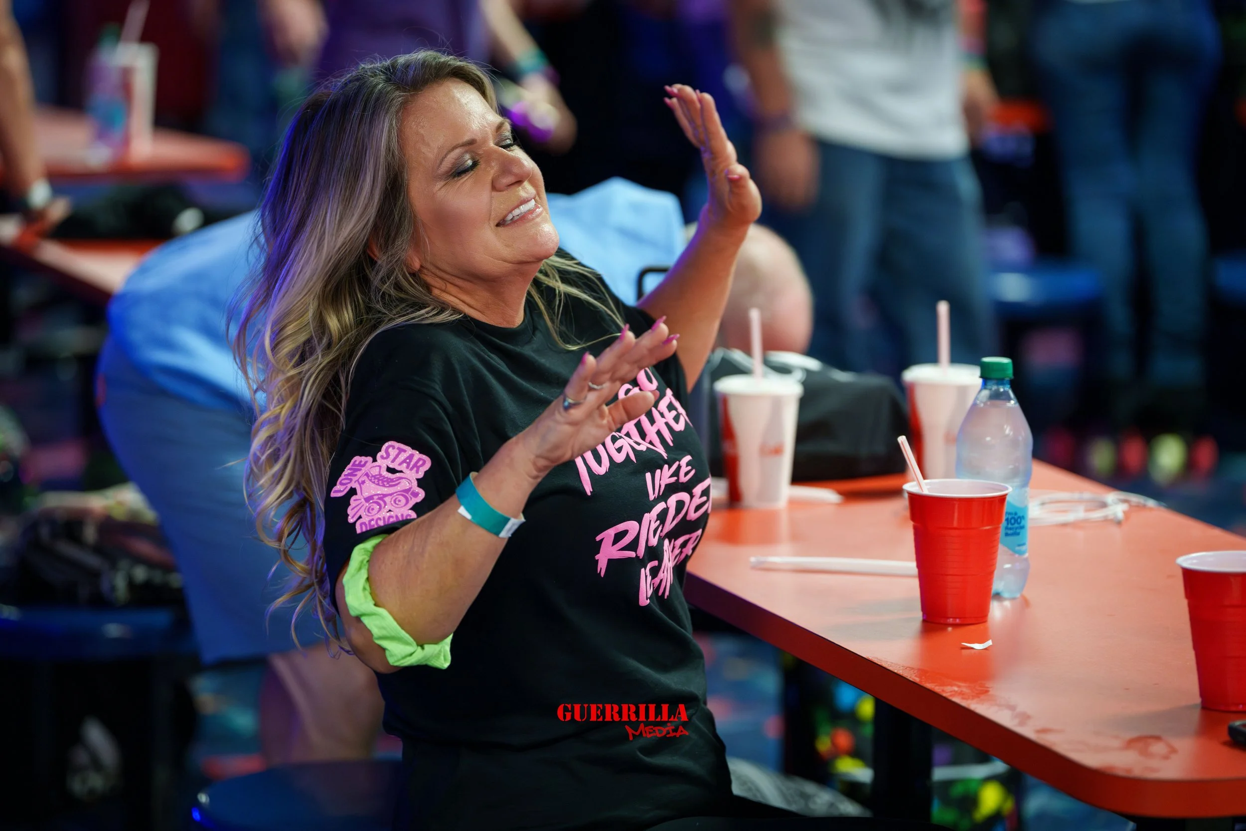 A woman with long wavy hair wearing a black T-shirt with pink text sitting at a bar or club, with her eyes closed and hands raised. There are red cups, a water bottle, and other people in the background.