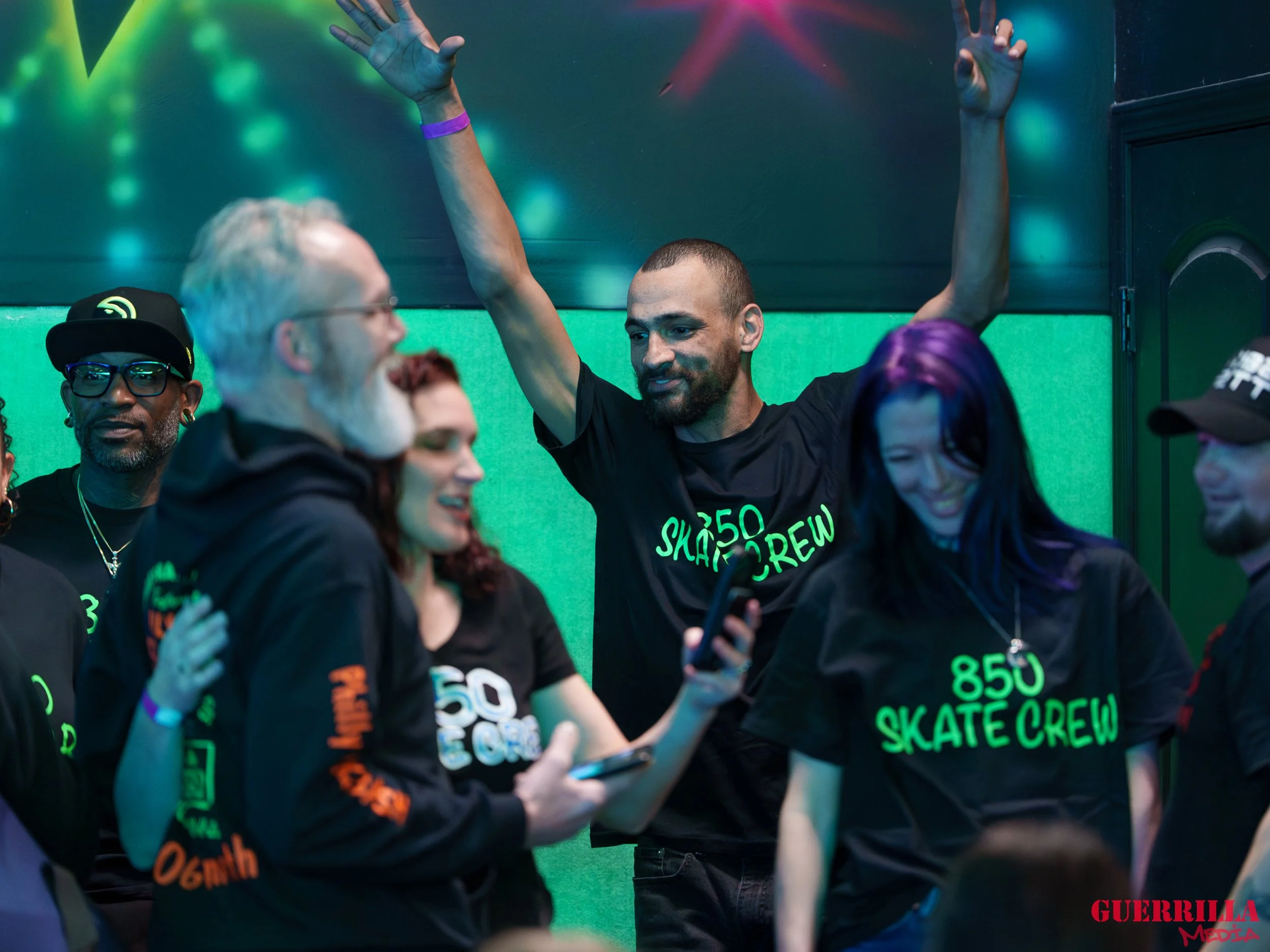 Group of people at a party or event, smiling and enjoying themselves. Some are wearing black shirts with green text, and one person has purple hair. There is a green background and colorful lighting.