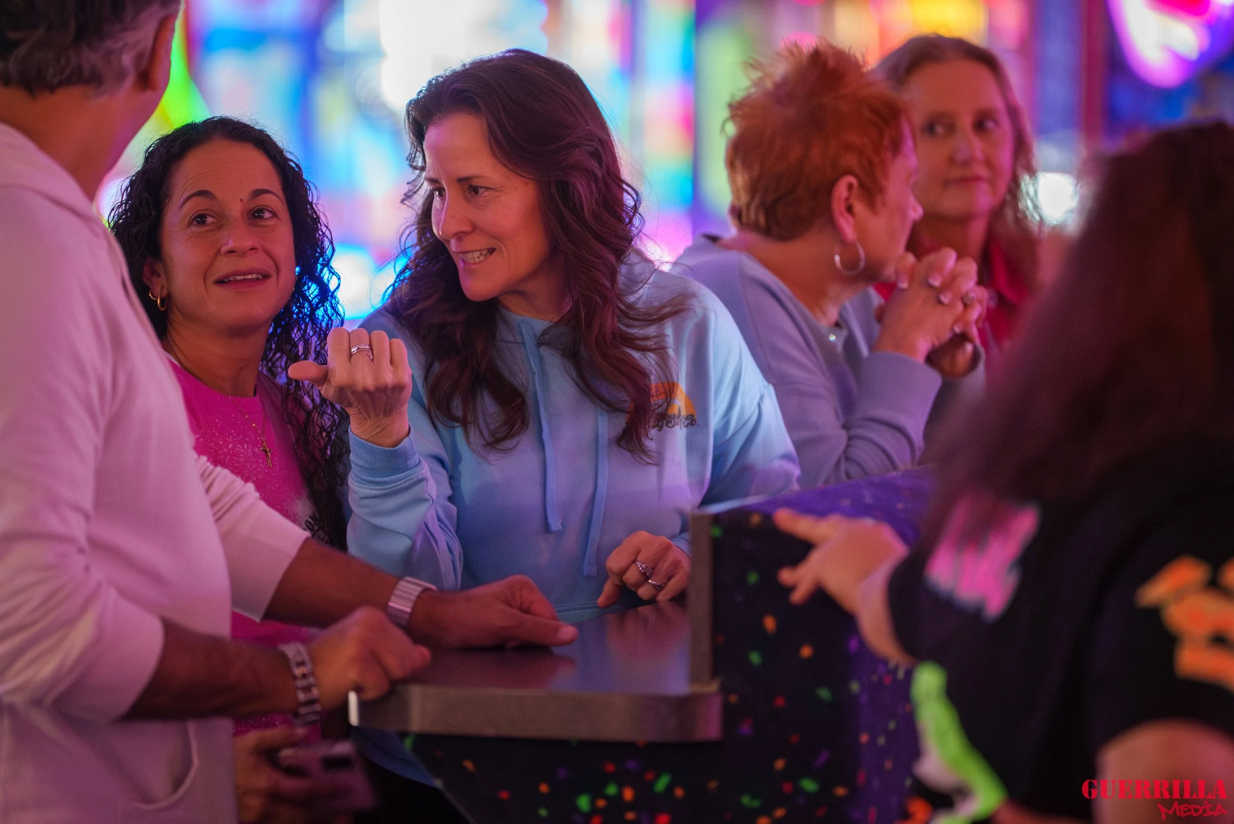 Group of women at a colorful, neon-lit bar or casino, engaged in conversation, with some smiling and pointing.