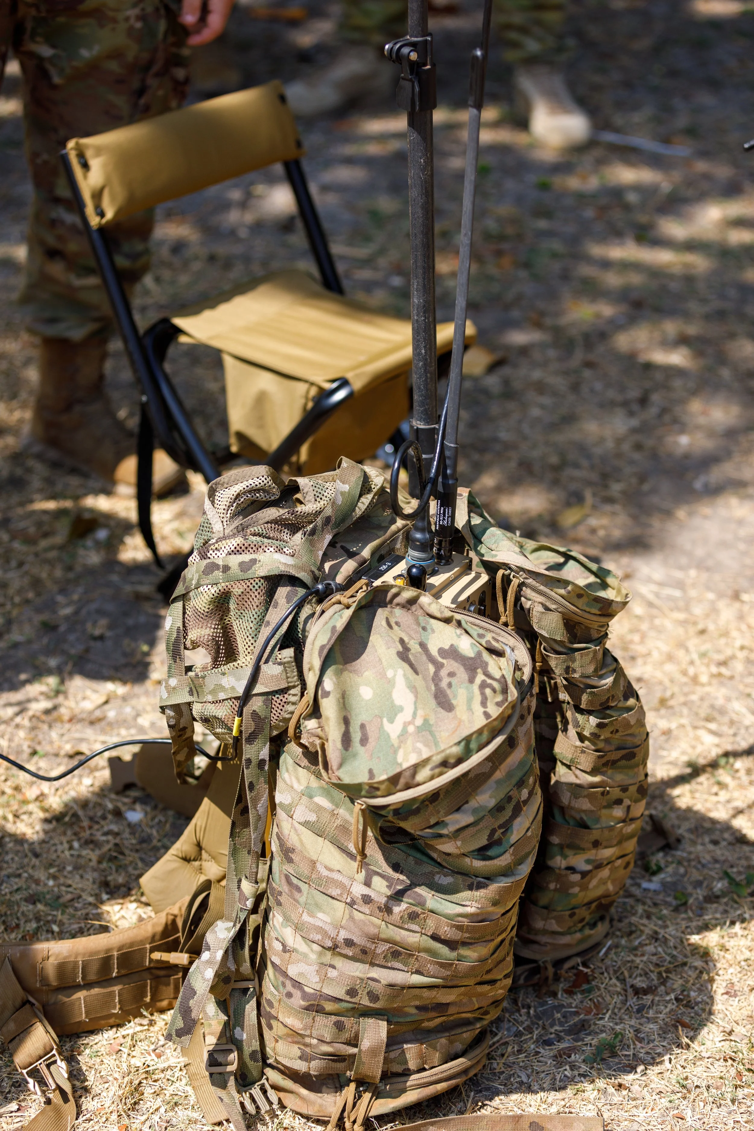Military camouflage tactical backpack with communication equipment and a small backpack on top, set on dirt ground; a tan folding chair and part of a person in camouflage pants are in background.
