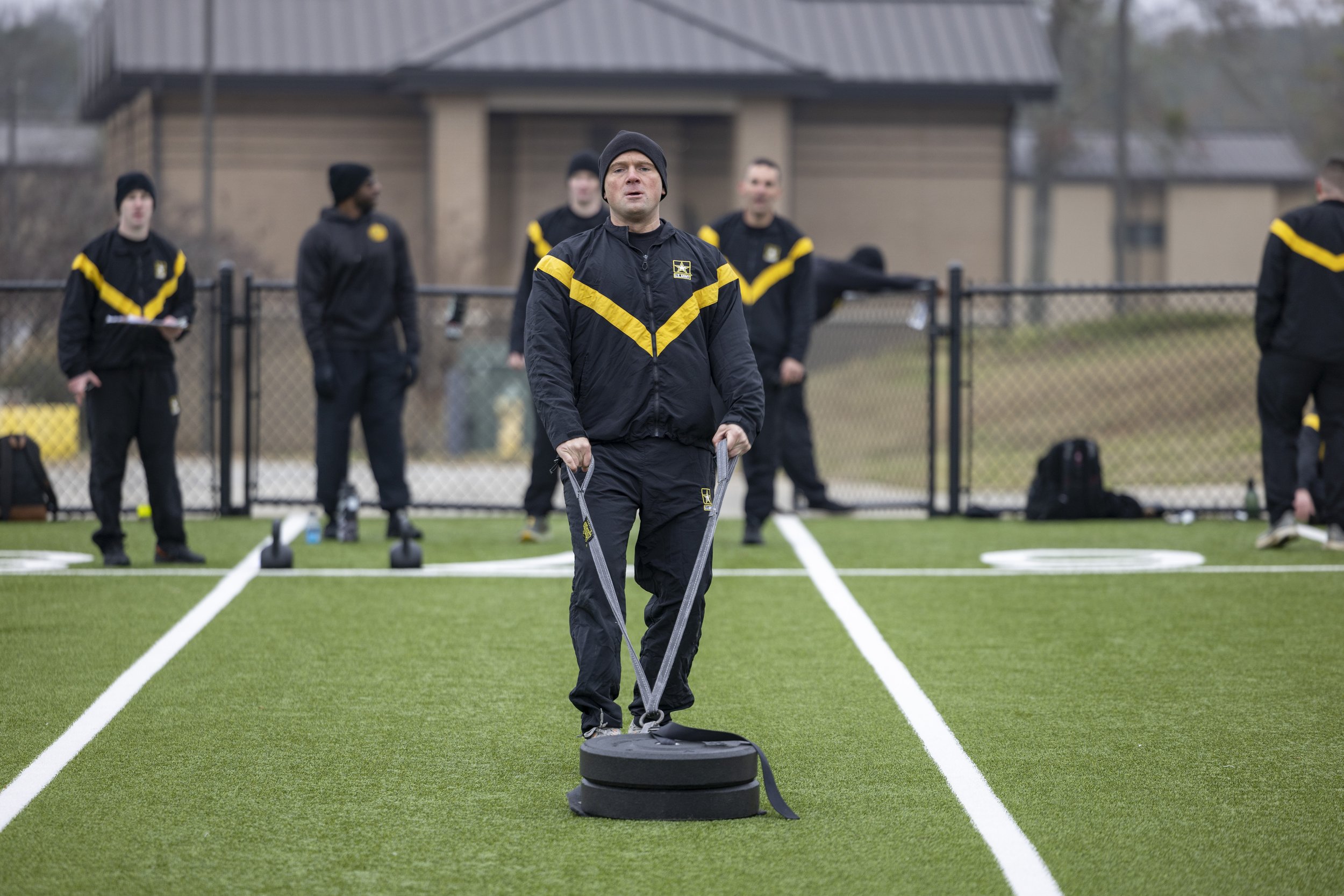 Man in black and yellow sportswear using a sled on a field with other men in similar sportswear in the background.