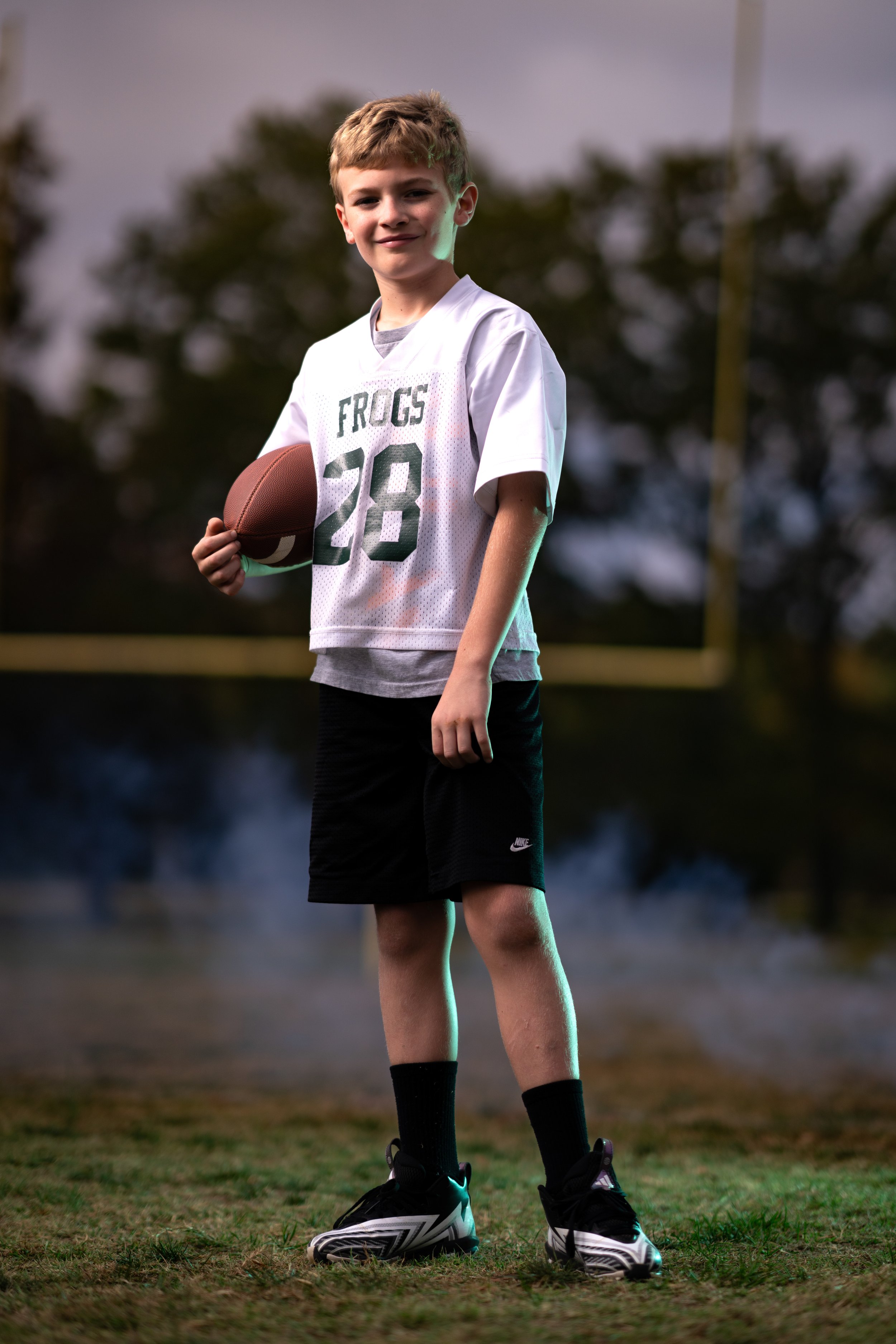 A young boy holding a football on the field during evening, wearing a white football jersey with the number 28 and the word "FROGS," black shorts, black socks, and black and white athletic shoes.