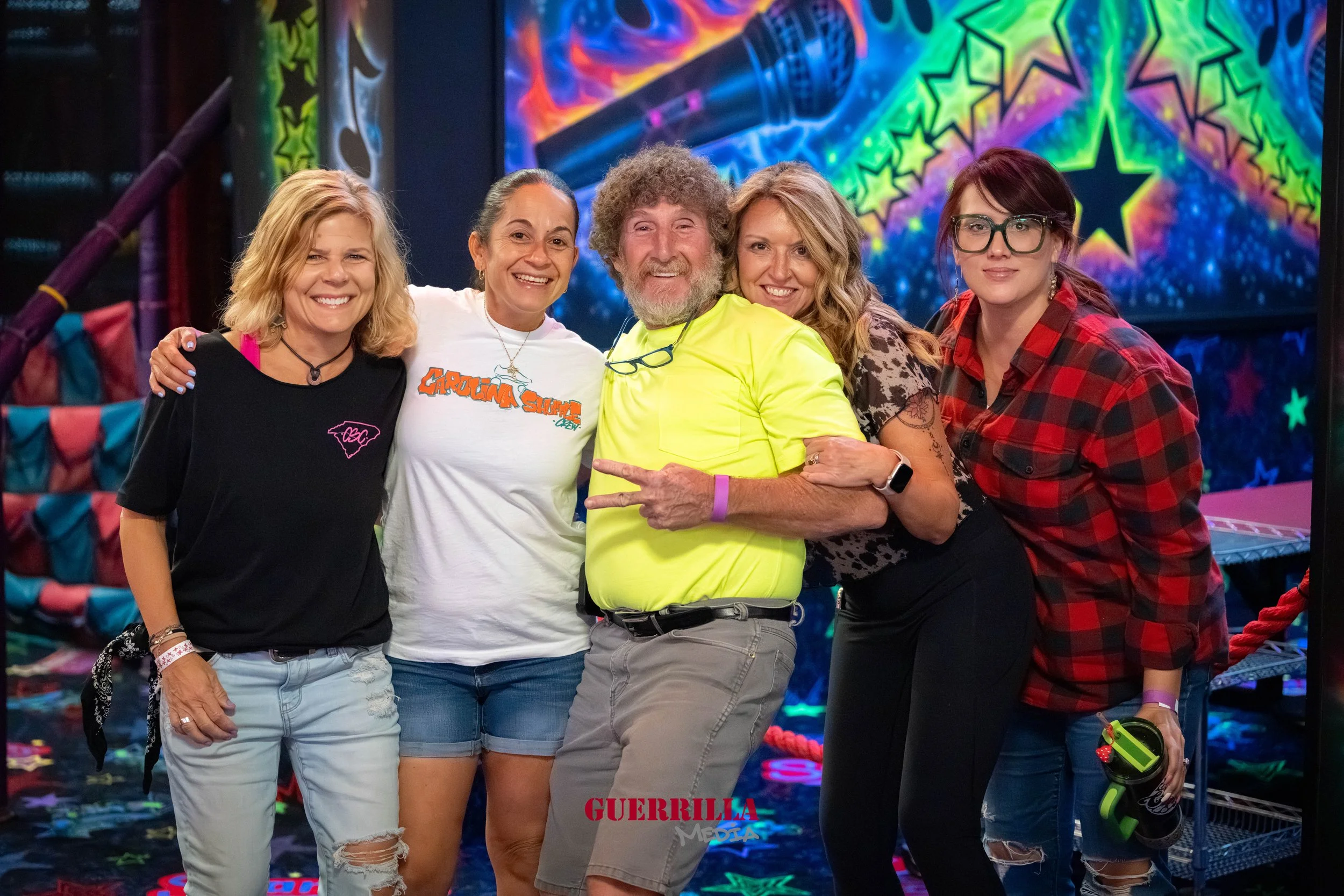 Five people standing together at an indoor amusement or arcade venue, smiling and posing for a photo. They are diverse in appearance and casual attire, with colorful, vibrant background lights and patterns.