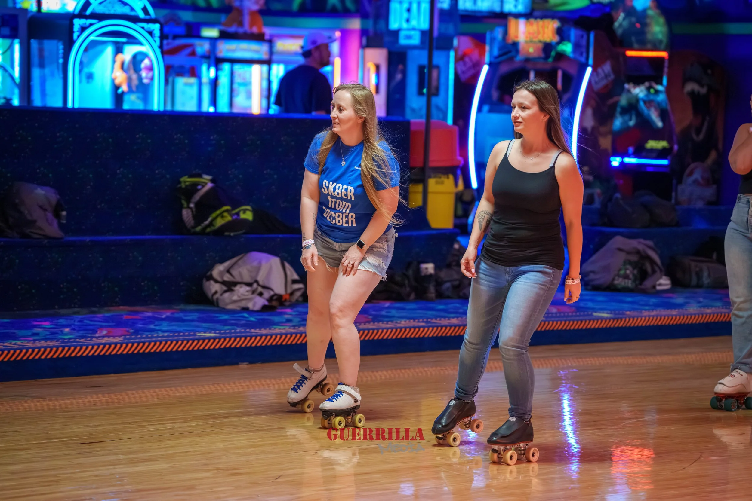 Two women roller skating indoors with neon lights and arcade games visible in the background.